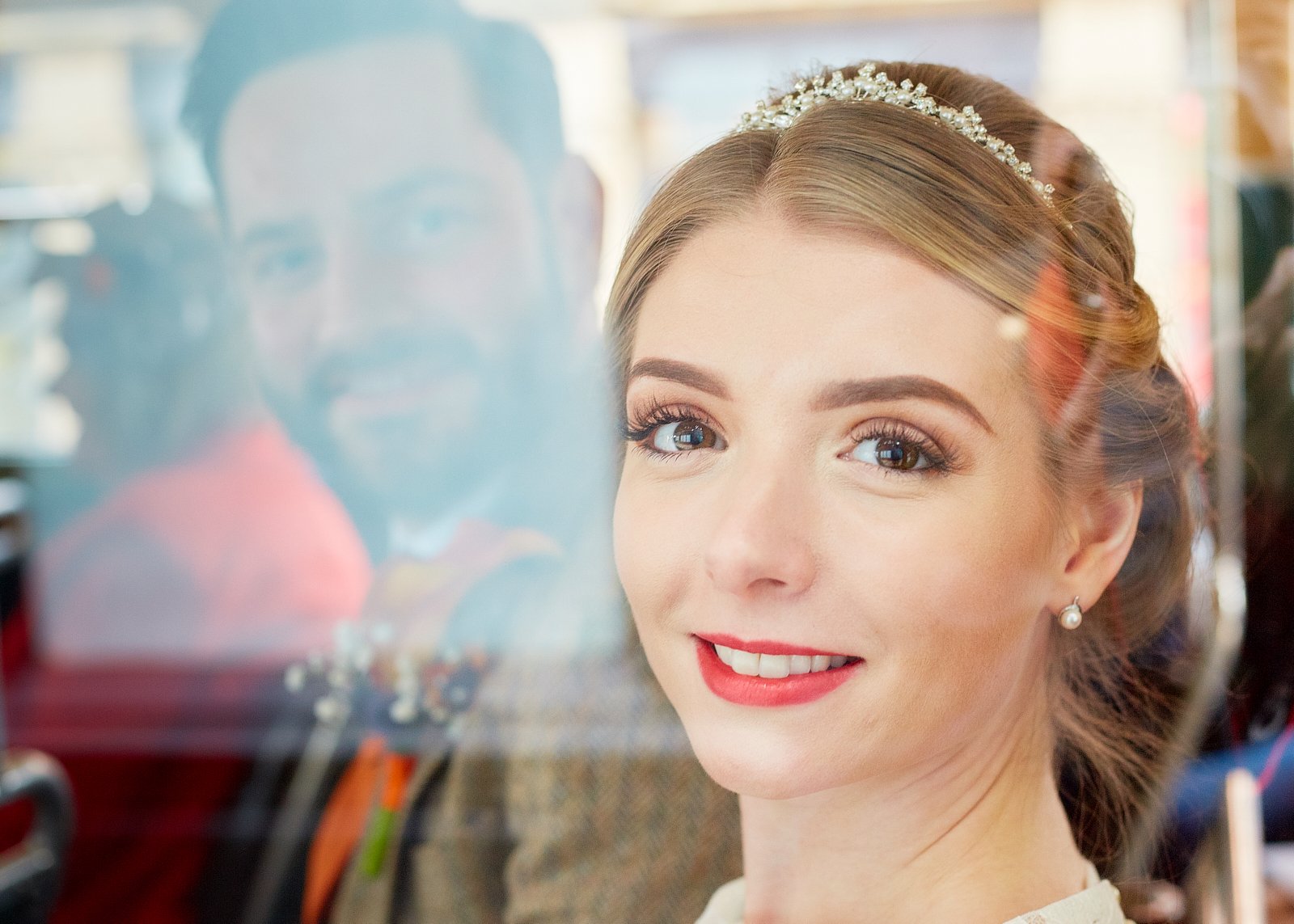 Close-up of a smiling bride with makeup and pearl earrings, looking through a glass reflection of a man with dark hair and a beard, in a bright indoor setting.