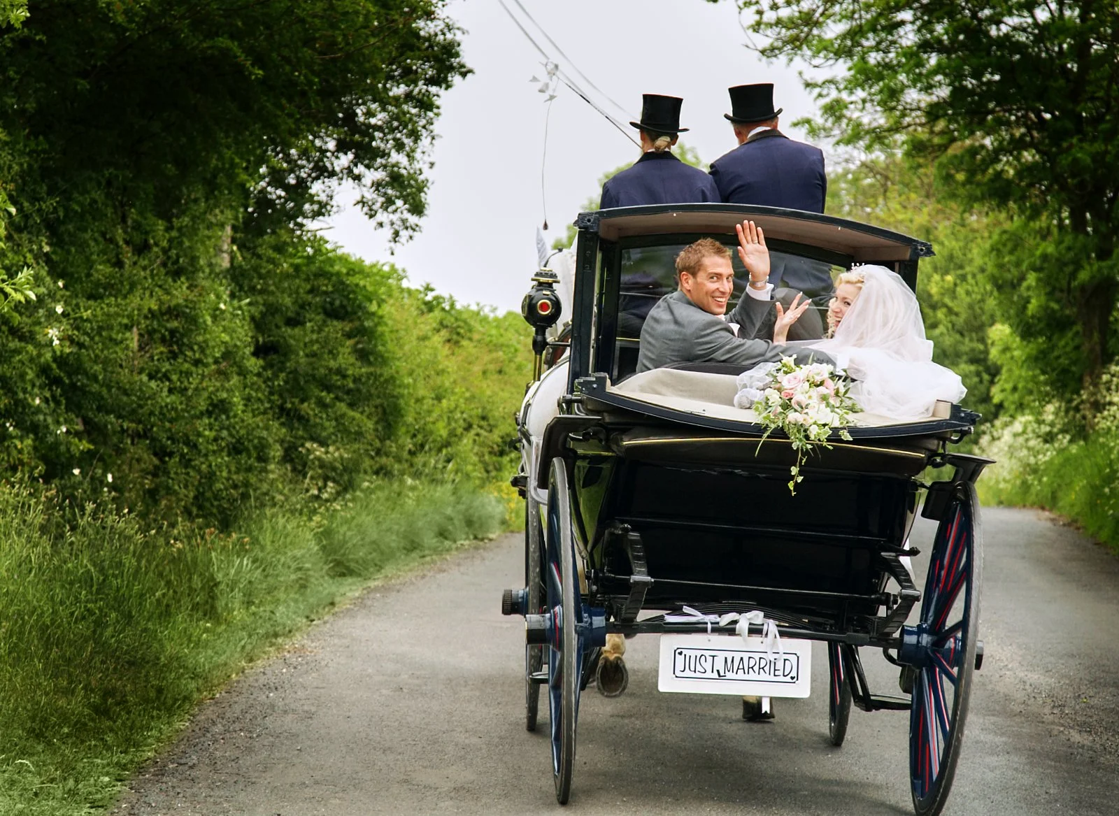 A newlywed couple riding in a horse-drawn carriage with a 'Just Married' sign on the back, waving and smiling, with two men in tuxedos and top hats sitting in the front, surrounded by green trees on a country road.