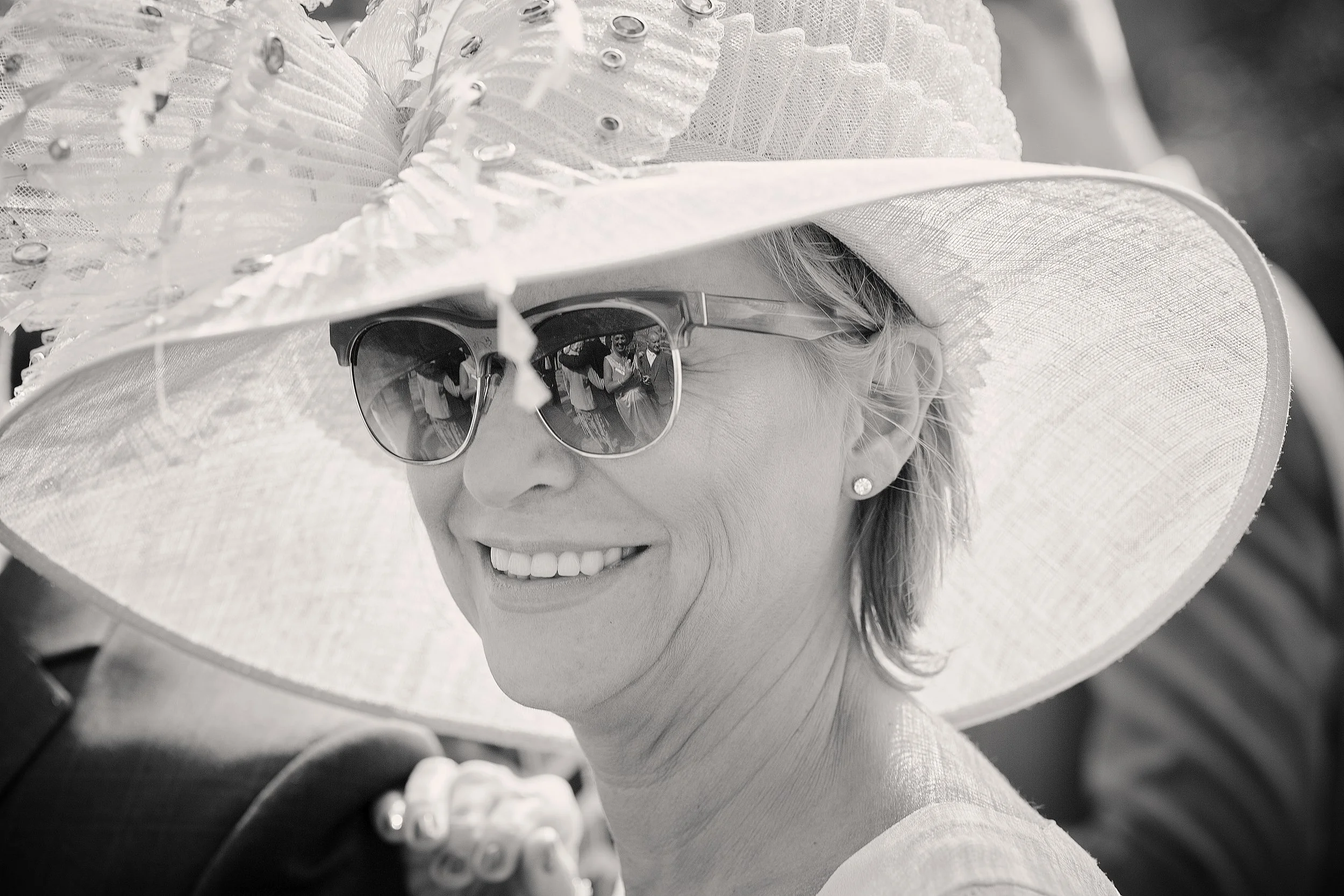 A woman wearing a large wide-brimmed hat with decorative details, sunglasses, and earrings, smiling.