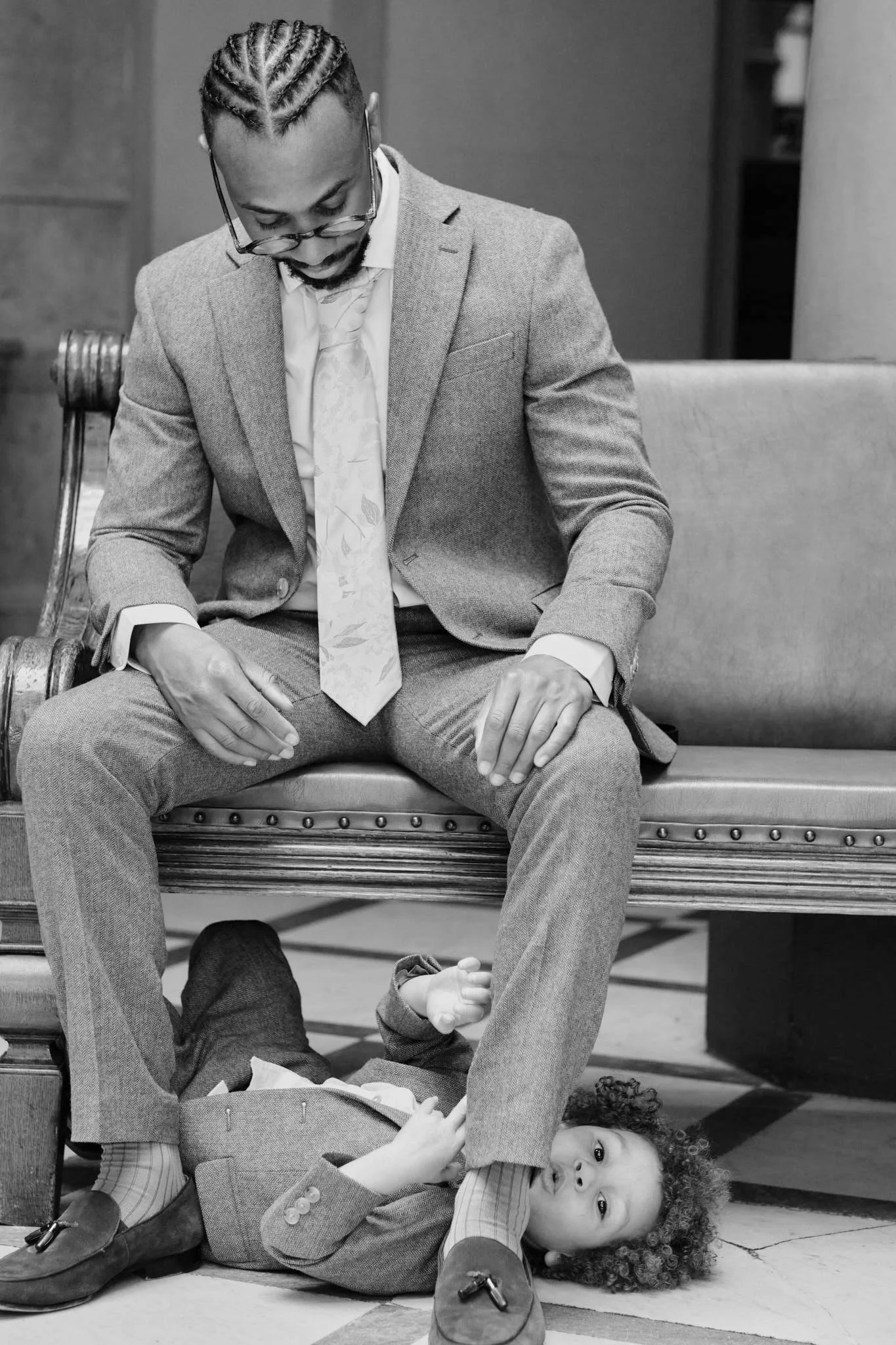 Black and white photo of a man in a suit sitting on a bench with his head bowed and hands on his lap, a young girl with curly hair lying on the floor beneath him, looking up at the camera with a relaxed expression.
