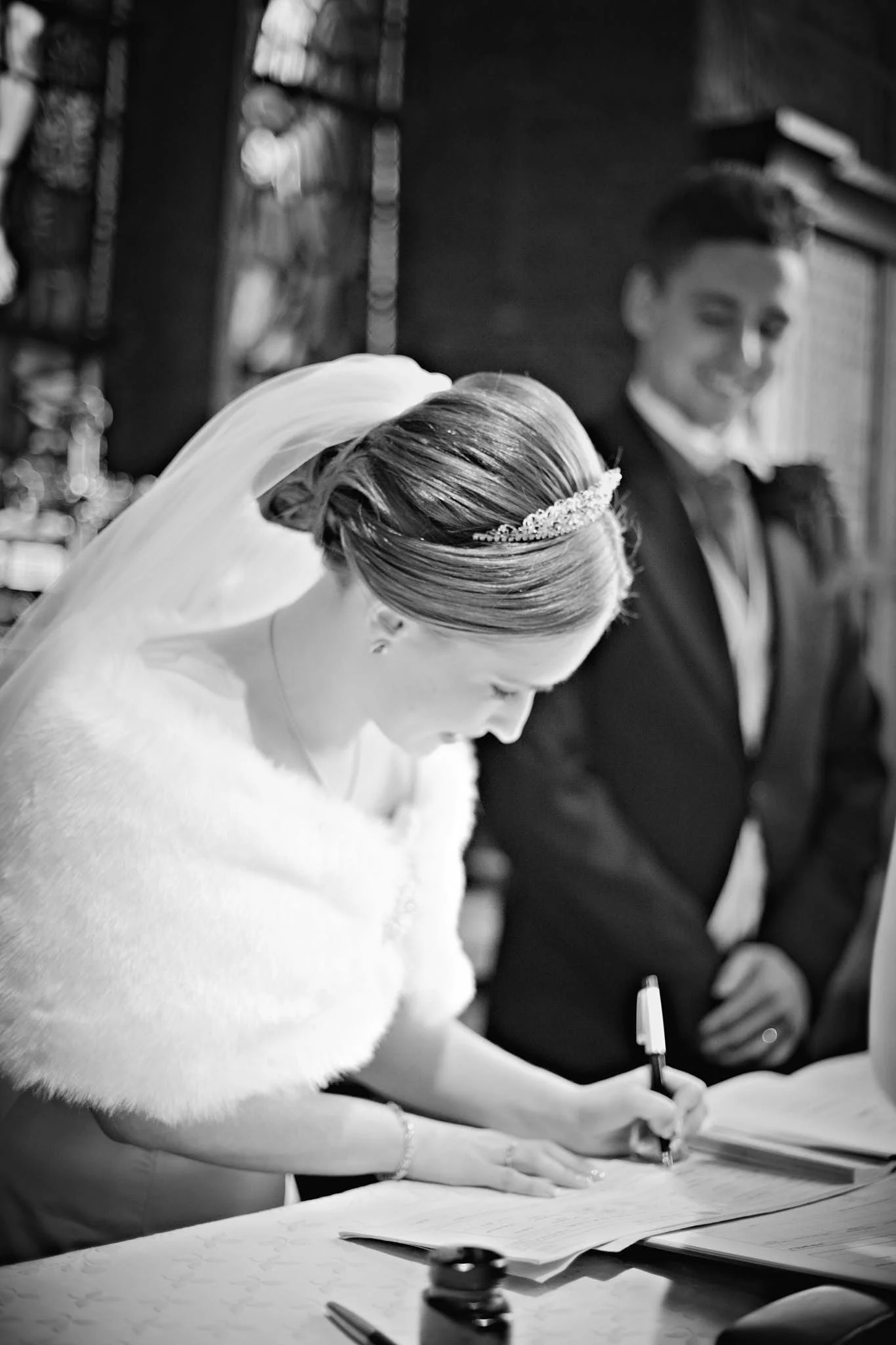 A woman with a wedding dress and veil signing a document at a table, with a man in a suit and tie standing behind her, smiling, in a room with bookshelves.