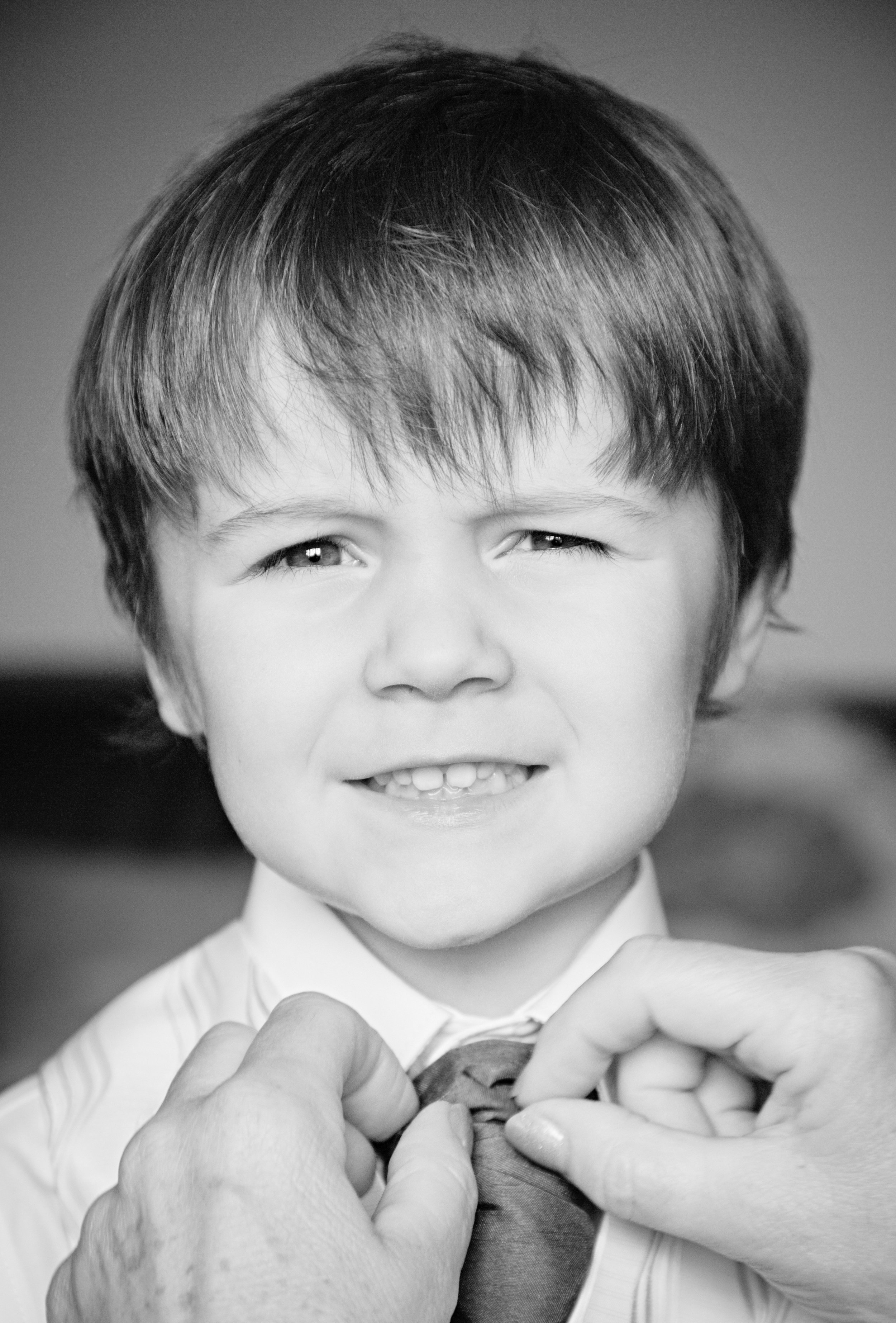 Black and white photo of a young boy smiling and winking, with a woman’s hands adjusting his tie in the foreground.