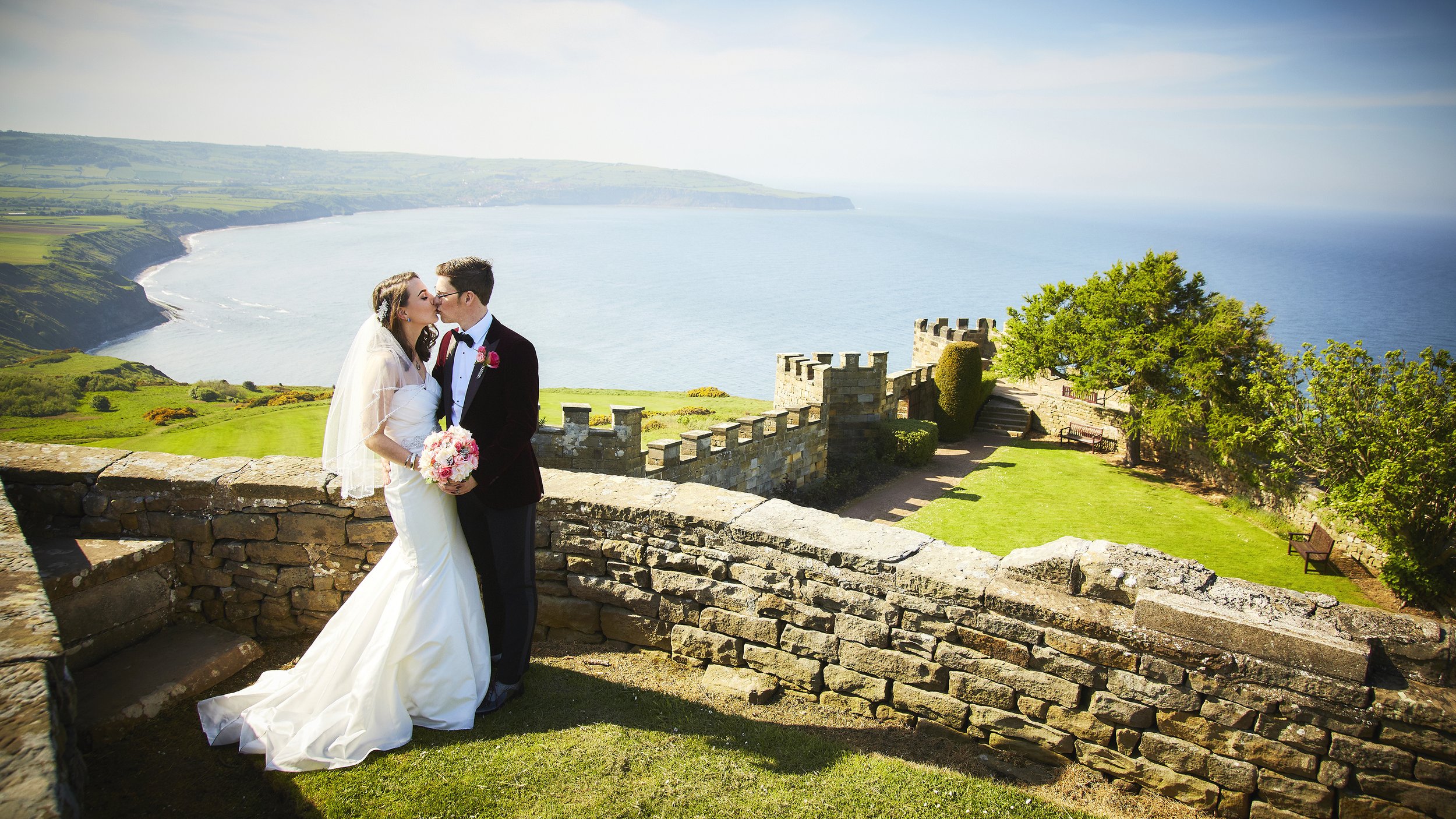 A bride and groom kissing on a stone balcony with a scenic coastal landscape in the background, including green hills and the ocean.