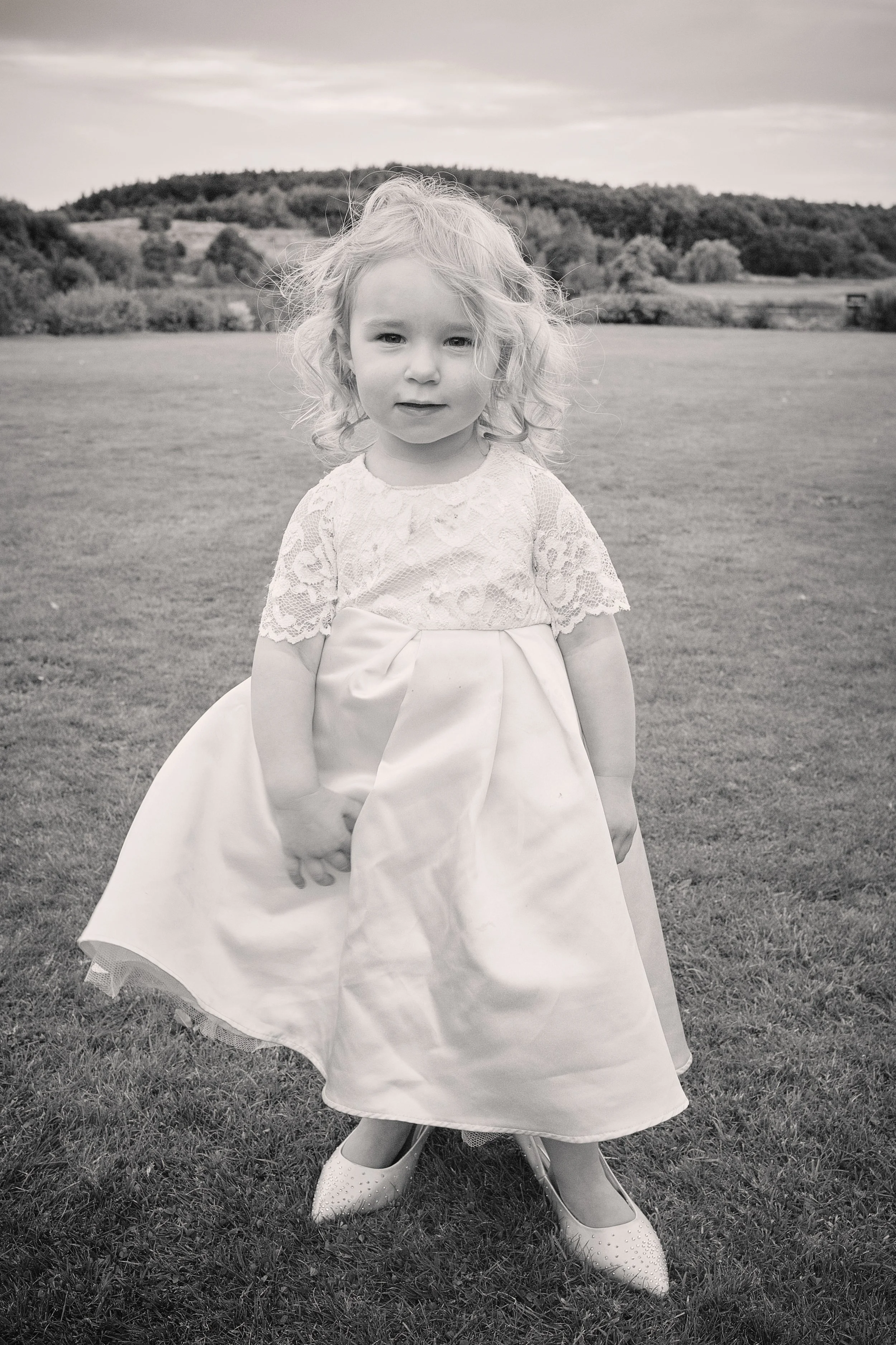 Black and white photo of a young girl in a fancy dress standing outdoors on the grass field, with hills and trees in the background. She is wearing lace top and a full skirt, along with heels. Her hair is curly and tousled.
