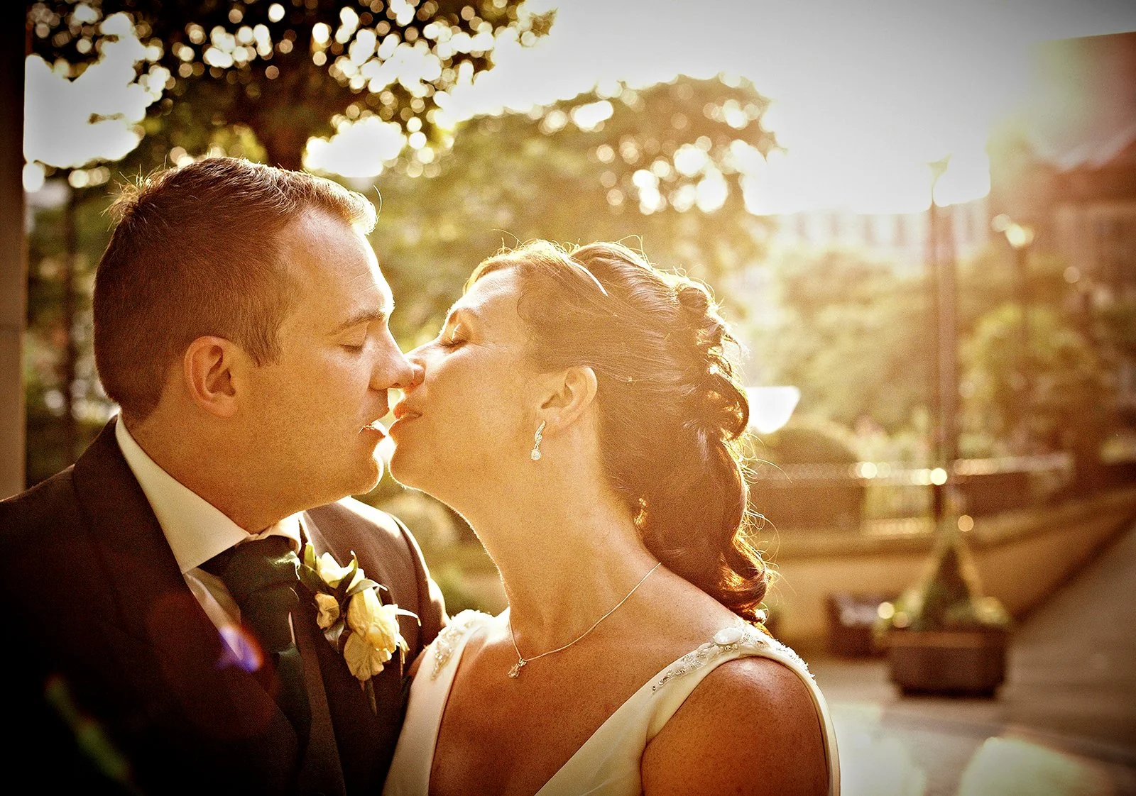 A couple sharing a kiss during their wedding outdoors at sunset, with trees and blurred background.