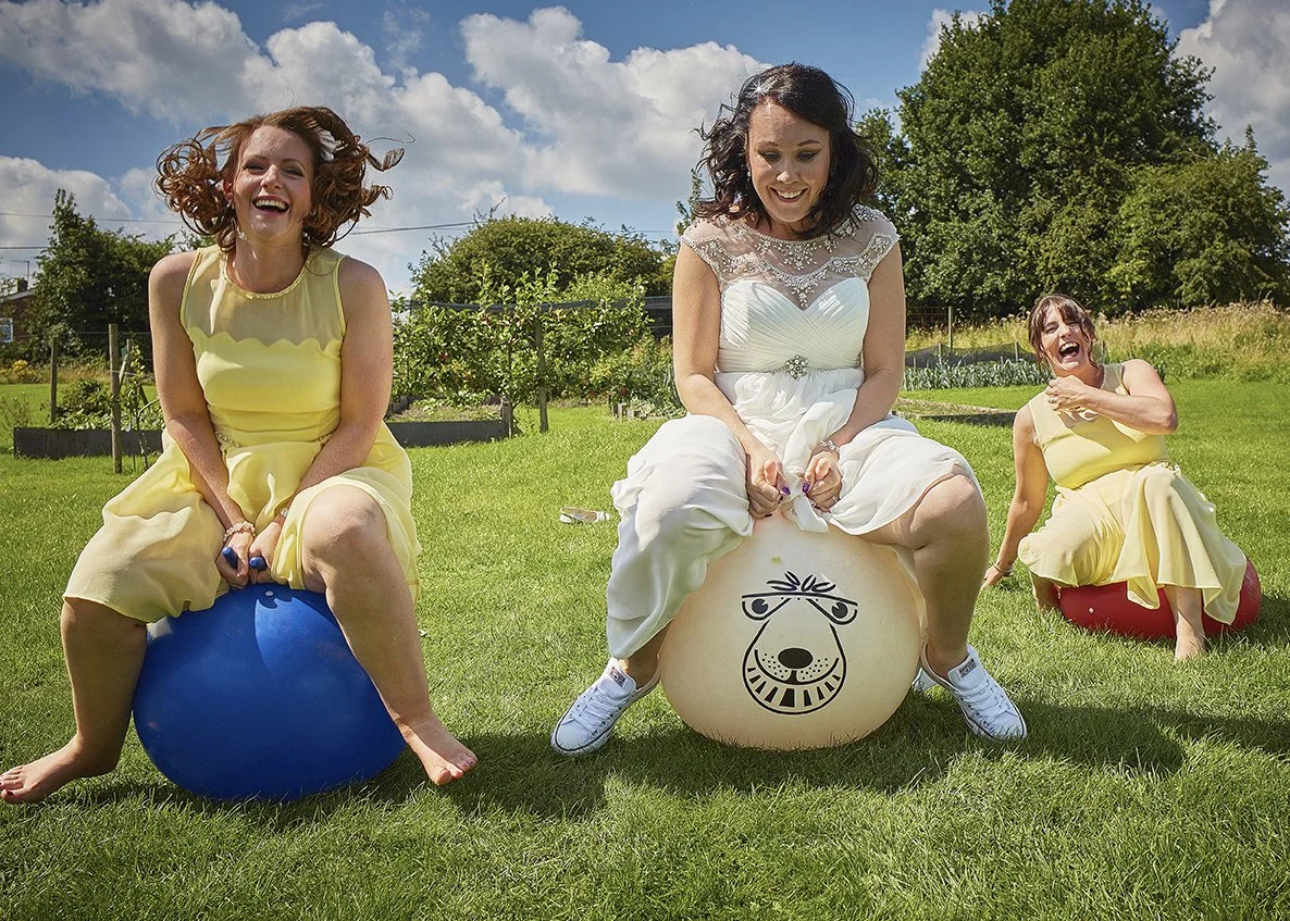 Three women in yellow dresses and a woman in a white dress playing on large colorful exercise balls outdoors on a sunny day with grassy field and trees