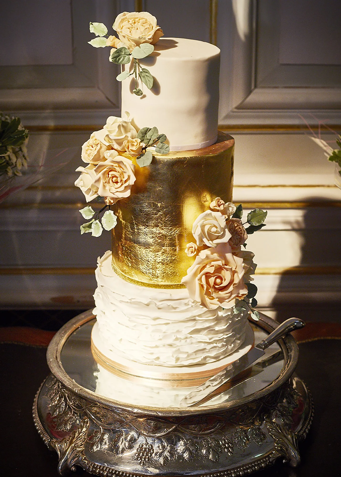 A three-tiered wedding cake decorated with white roses and green leaves, with a gold-painted middle tier, presented on an ornate silver tray.