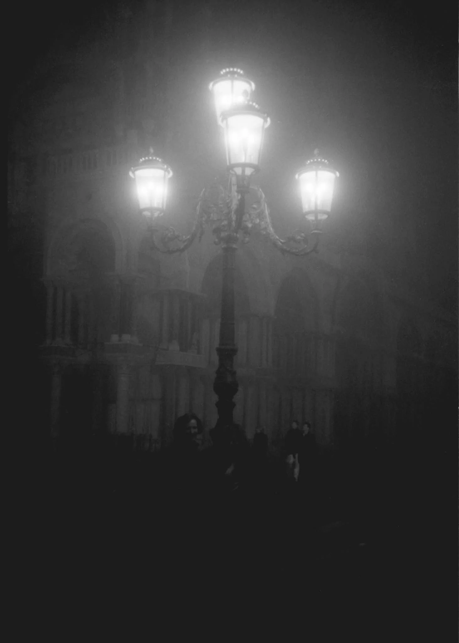 A vintage-style streetlamp with three glowing lanterns in a foggy night setting. The streetlamp and surrounding area have a slightly mysterious, eerie atmosphere, with silhouettes of people at the bottom of the image.