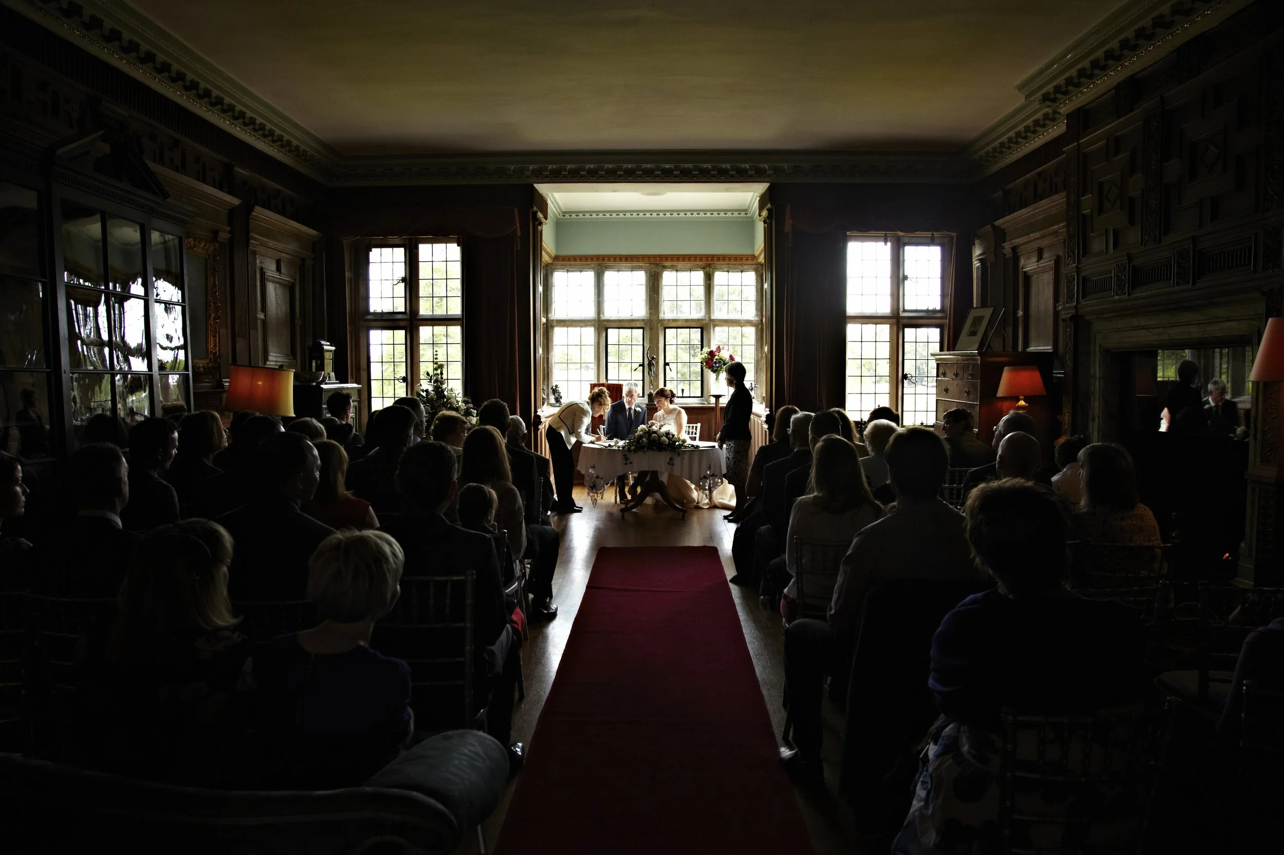 A wedding ceremony taking place in a dimly lit, historic room with large windows and wooden paneling. The bride and groom are seated at a table with floral arrangements, while a person stands nearby. Guests are seated facing the couple, and the room 