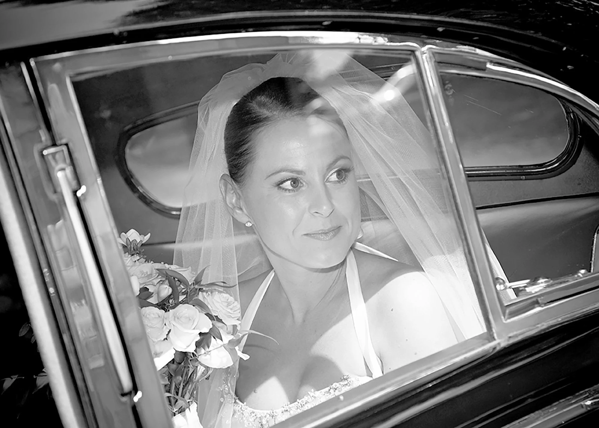 Black and white photo of a bride with dark hair in a bun, wearing a veil and holding a bouquet of roses, visible through the window of a vehicle.