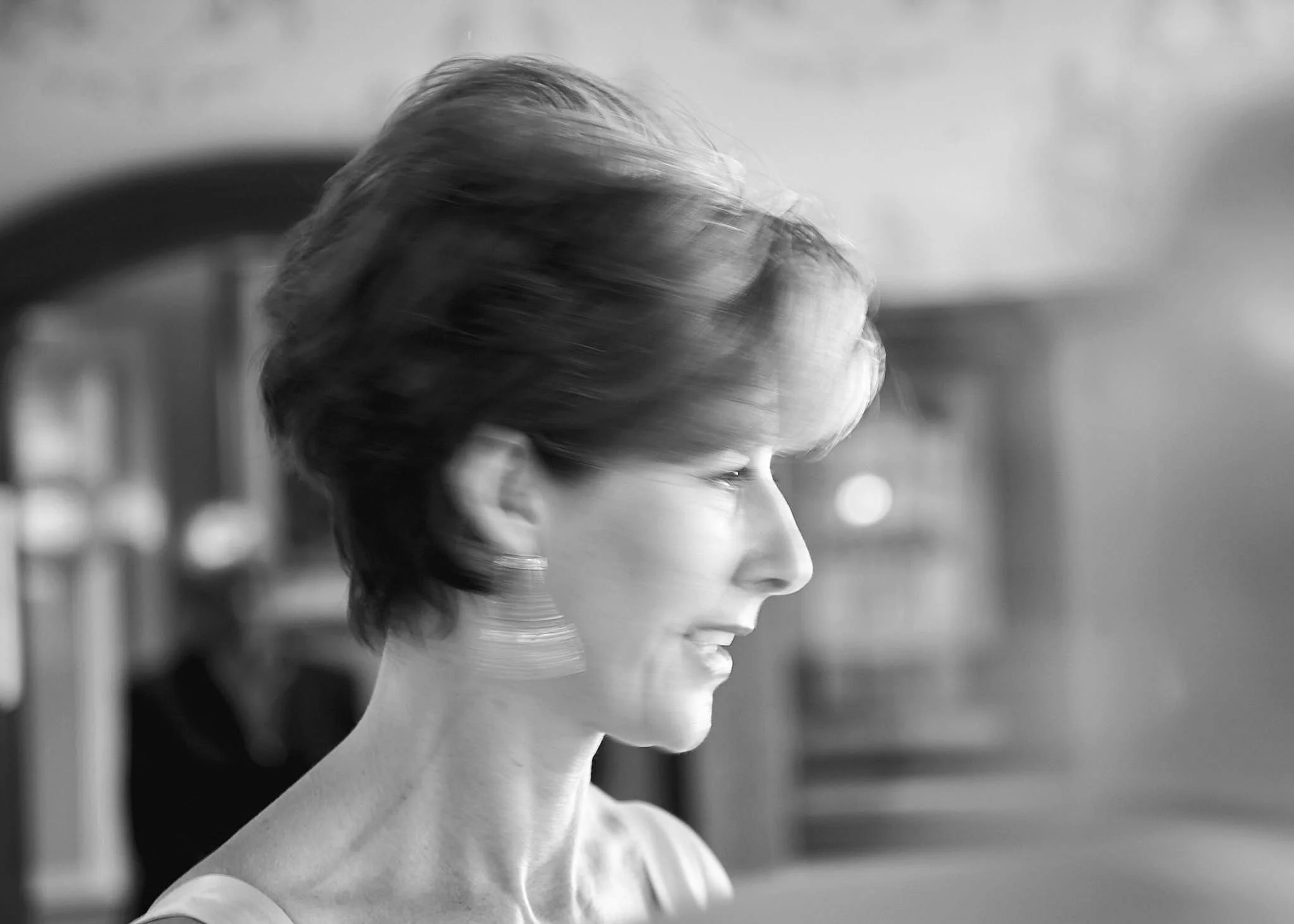 A black-and-white profile photo of a woman with short, wavy hair, looking down with a pensive expression, with blurred background.