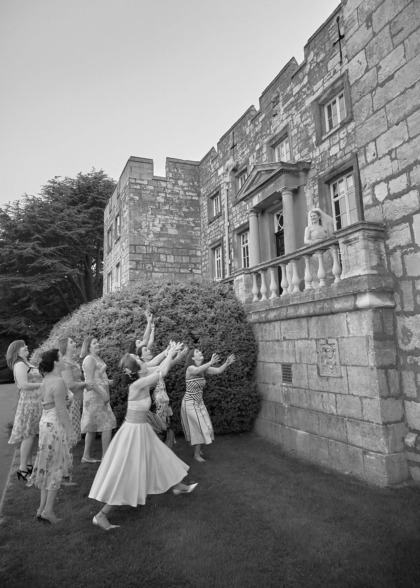 A group of women in summer dresses are happily running and reaching towards a smiling woman on a balcony of a large stone building. The building has multiple windows and a columned entrance, with a bush and trees in the background.