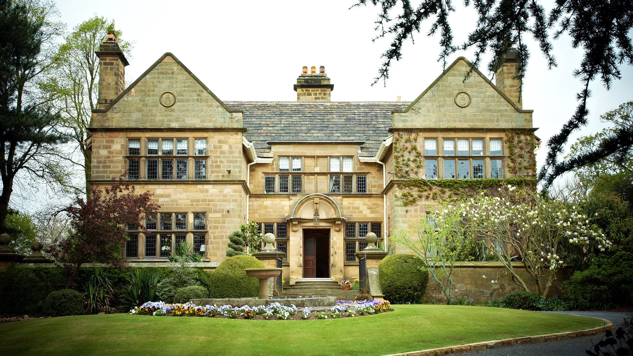 A large, historic stone house with multiple gables, large windows, and a central entrance, surrounded by a well-maintained garden with trees, bushes, and flowering plants.