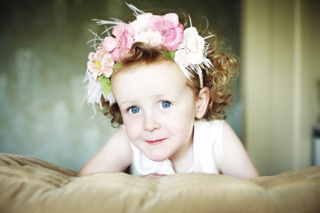 A young girl with curly red hair and blue eyes wearing a floral headband with pink and white flowers looks directly at the camera.