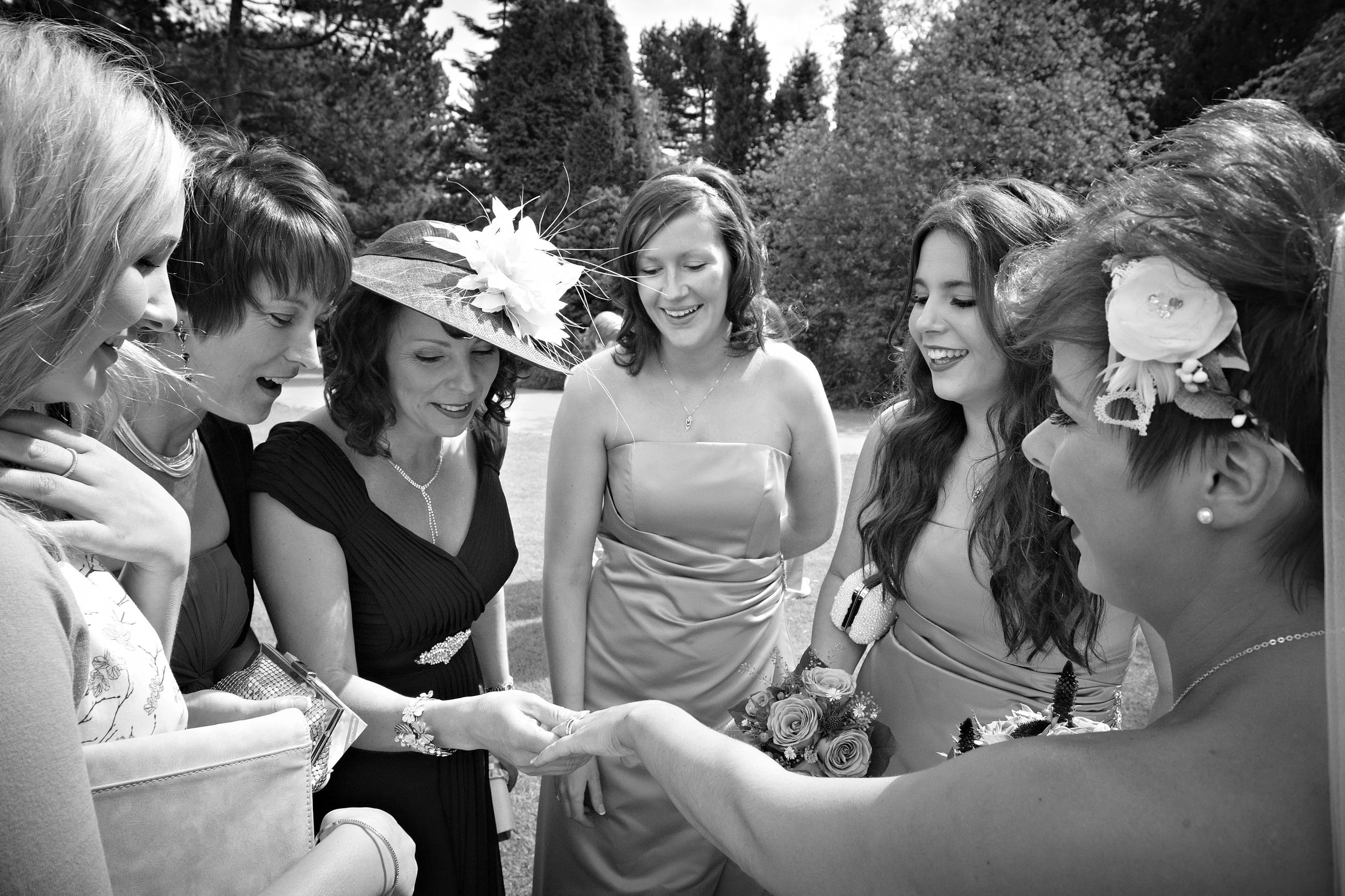 Black and white photo of seven women, gathered outdoors, smiling and looking down, with some holding flowers.