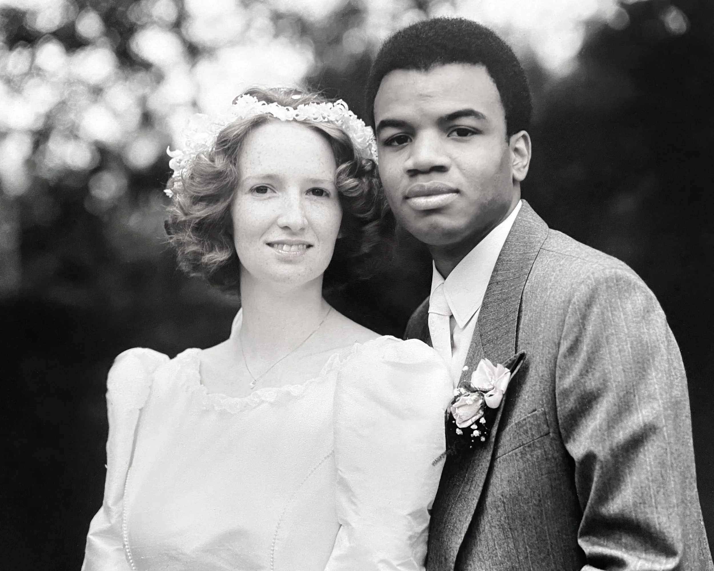 A black and white photograph of a young couple on their wedding day, with the bride wearing a lace dress and veil, and the groom dressed in a suit with a boutonniere. They are standing close together outdoors with a blurred background of trees or foliage.