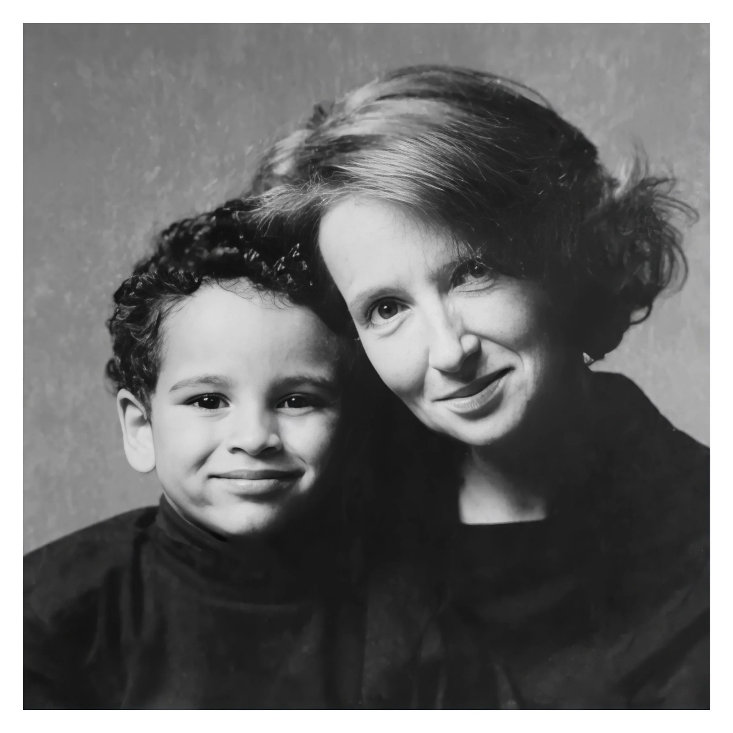 Black and white photograph of a woman and a young girl smiling, close-up of their faces, woman's hair and the girl's face prominently featured.