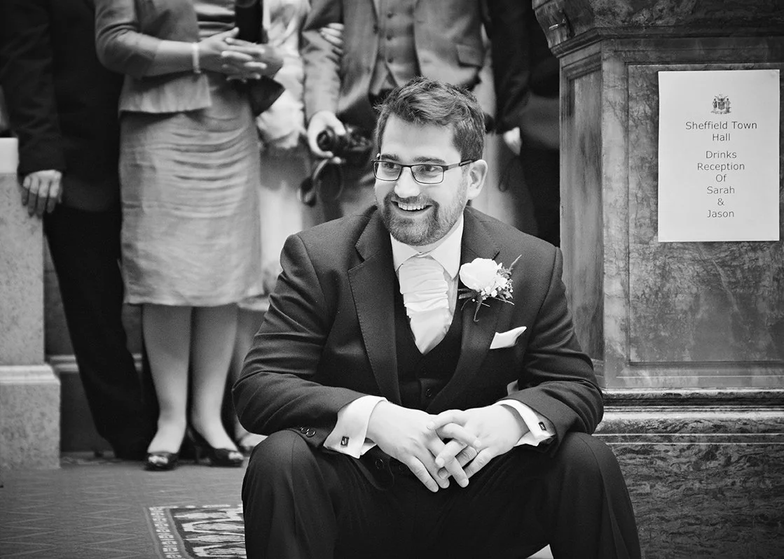 A happy man in a suit with a boutonniere, sitting on the floor at a wedding reception, with guests in the background and a sign on the wall reading 'Sheffield Town Hall, Drinks Reception, Of Sarah & Jason.'