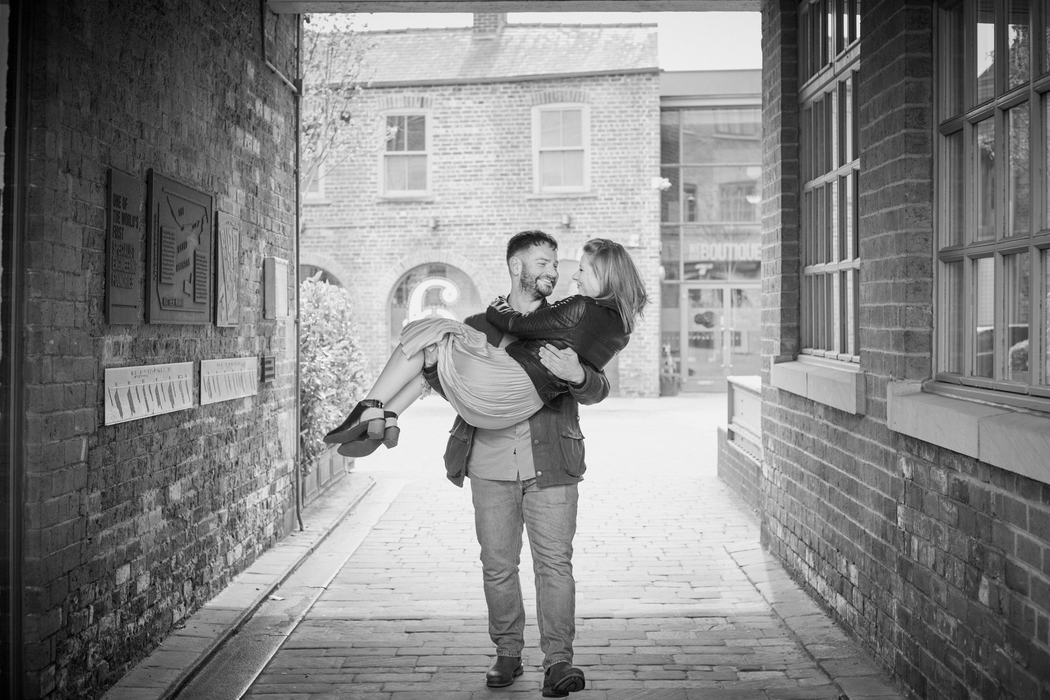 A man carrying a woman in a lively alleyway between brick buildings, holding her in his arms, both smiling and engaging affectionately.