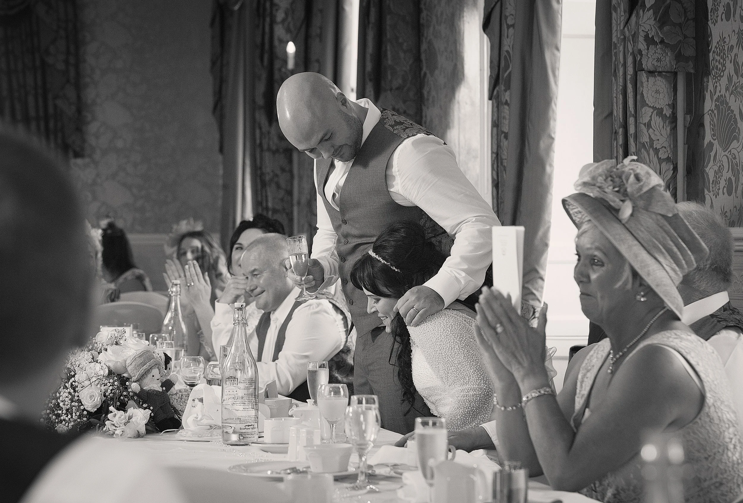 A black and white photo of a man serving food during a banquet, with guests seated at a long table with wine glasses, floral centerpieces, and table settings. The man is dressed in formal attire, and the atmosphere appears elegant and celebratory.