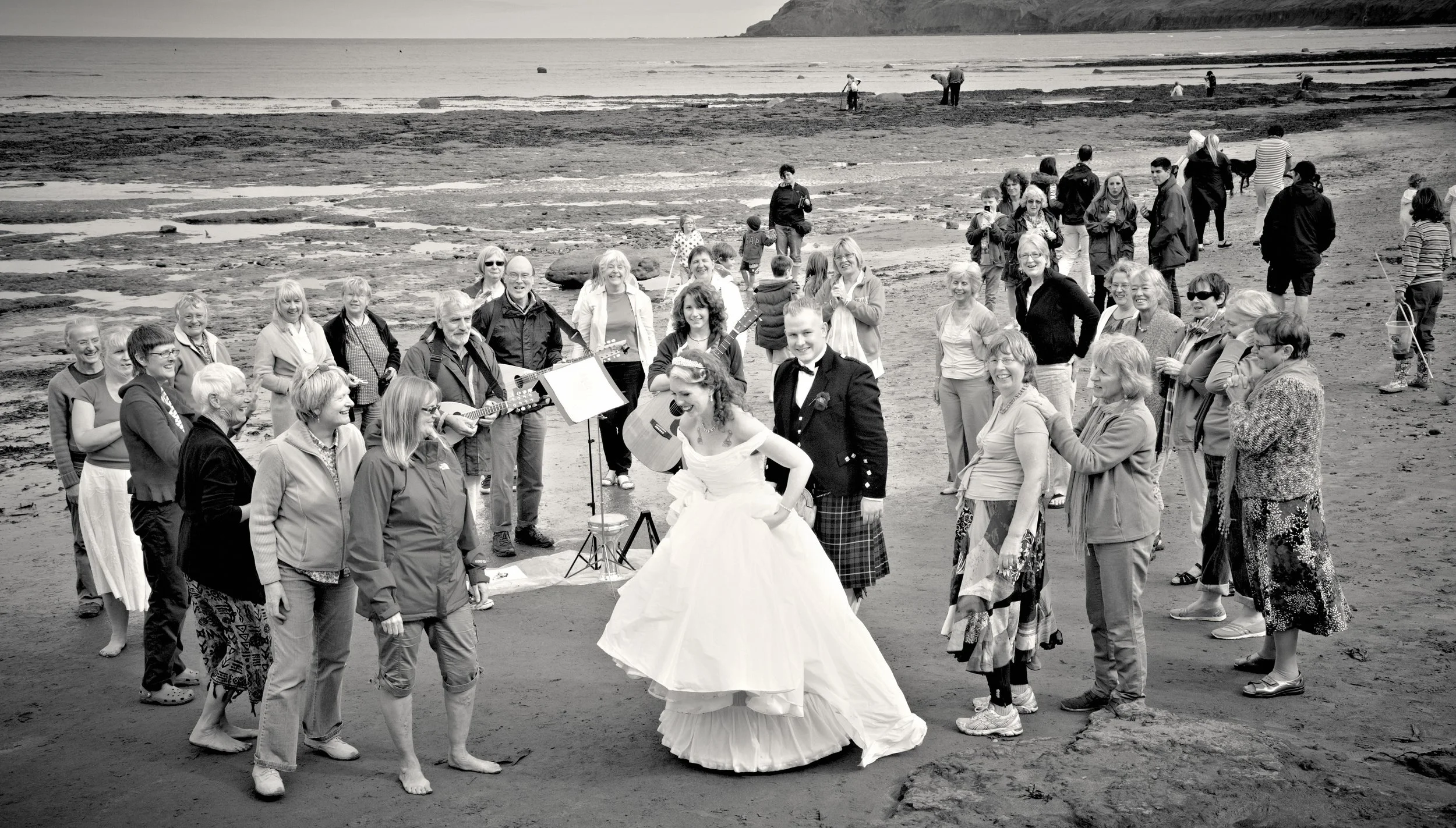 A group of people gathered on a beach, some playing guitars and others smiling, with a woman in a white wedding dress and a man in a suit at the center, indicating a wedding celebration.
