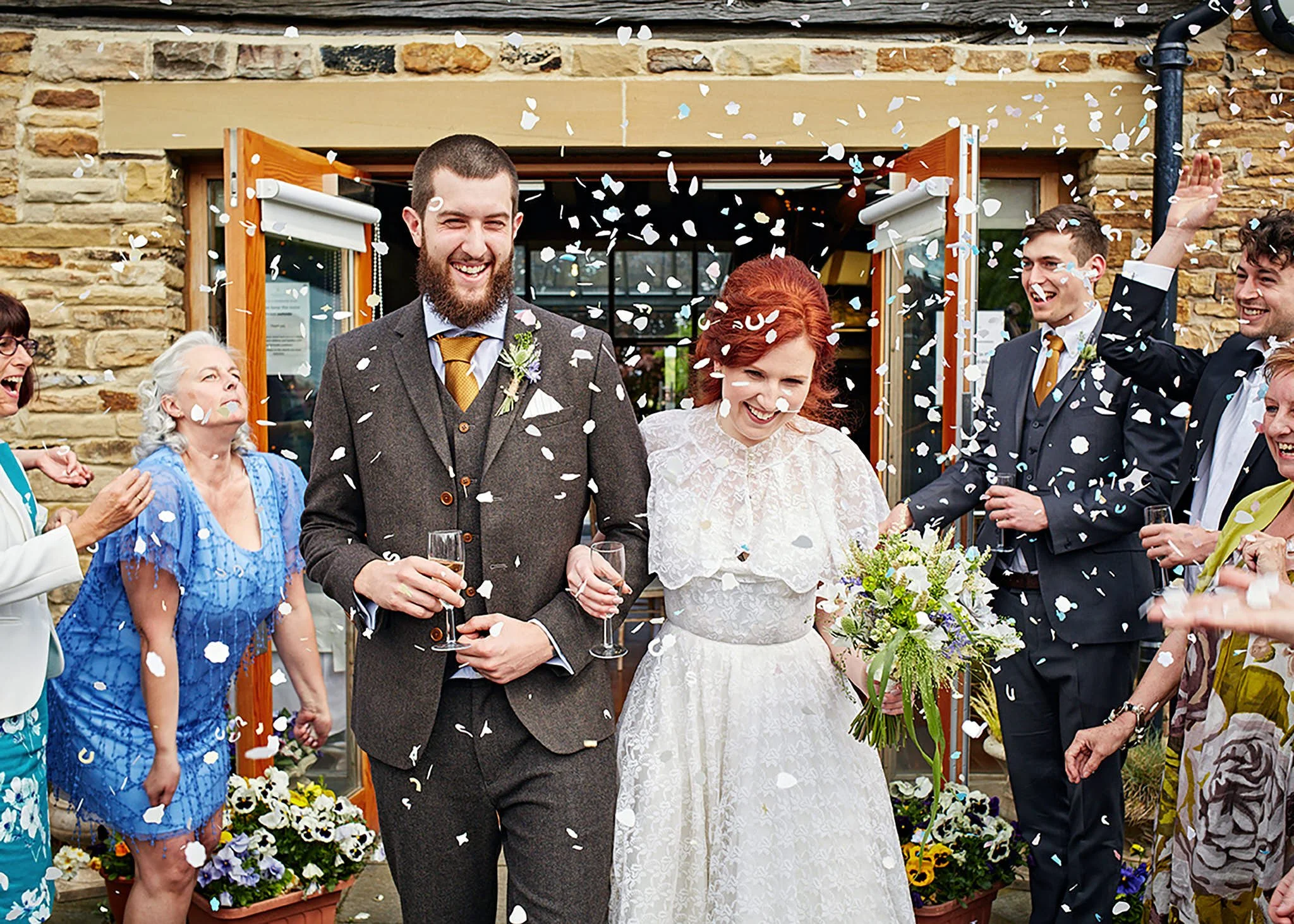 A wedding celebration with a bride and groom standing outside in front of a stone building, surrounded by family and friends, with white confetti falling around them.