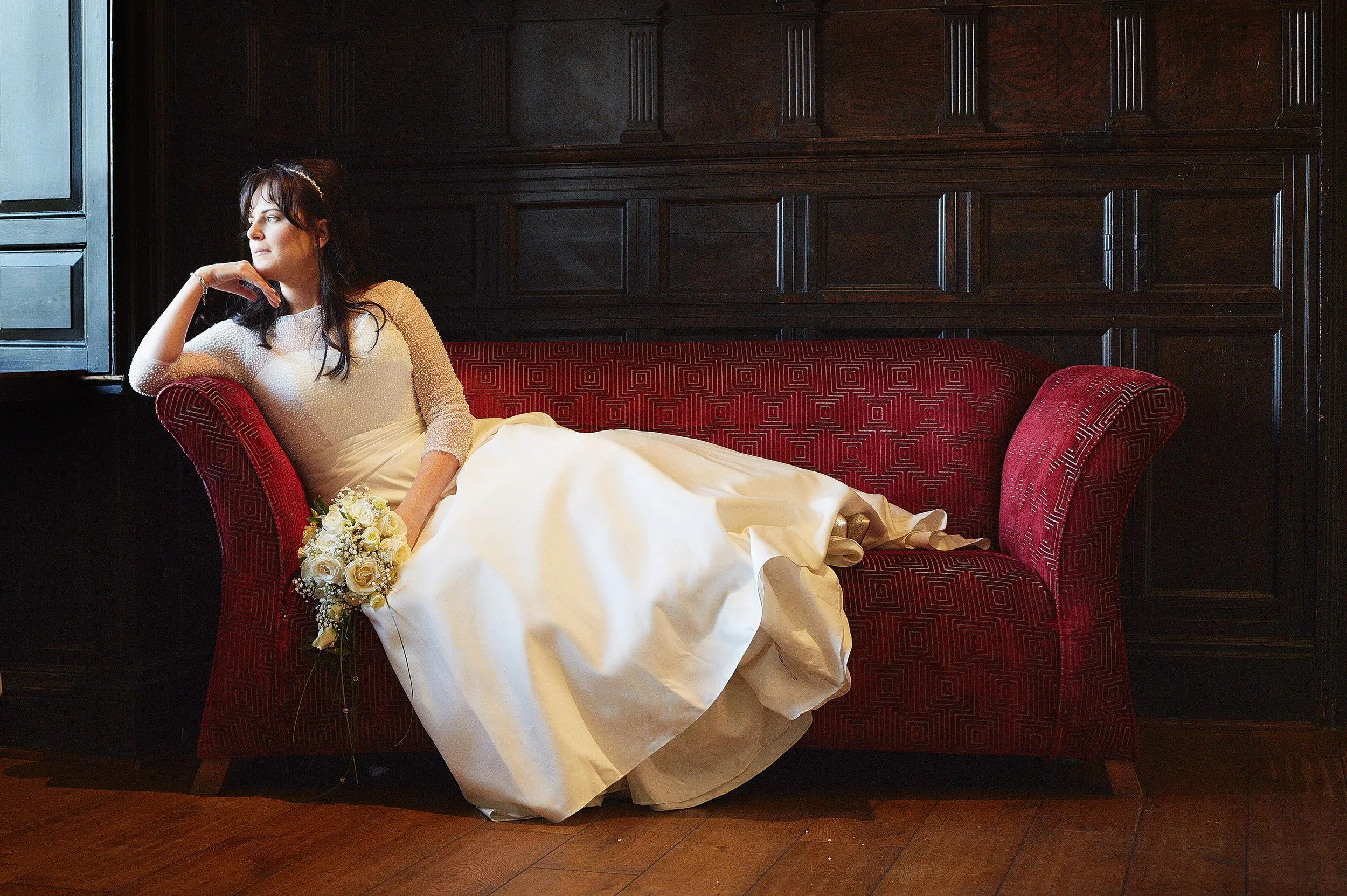 A woman in a white wedding dress reclining on a red-patterned vintage-style sofa in a dark wood-paneled room, looking out the window with a thoughtful expression, holding a bouquet of white roses.