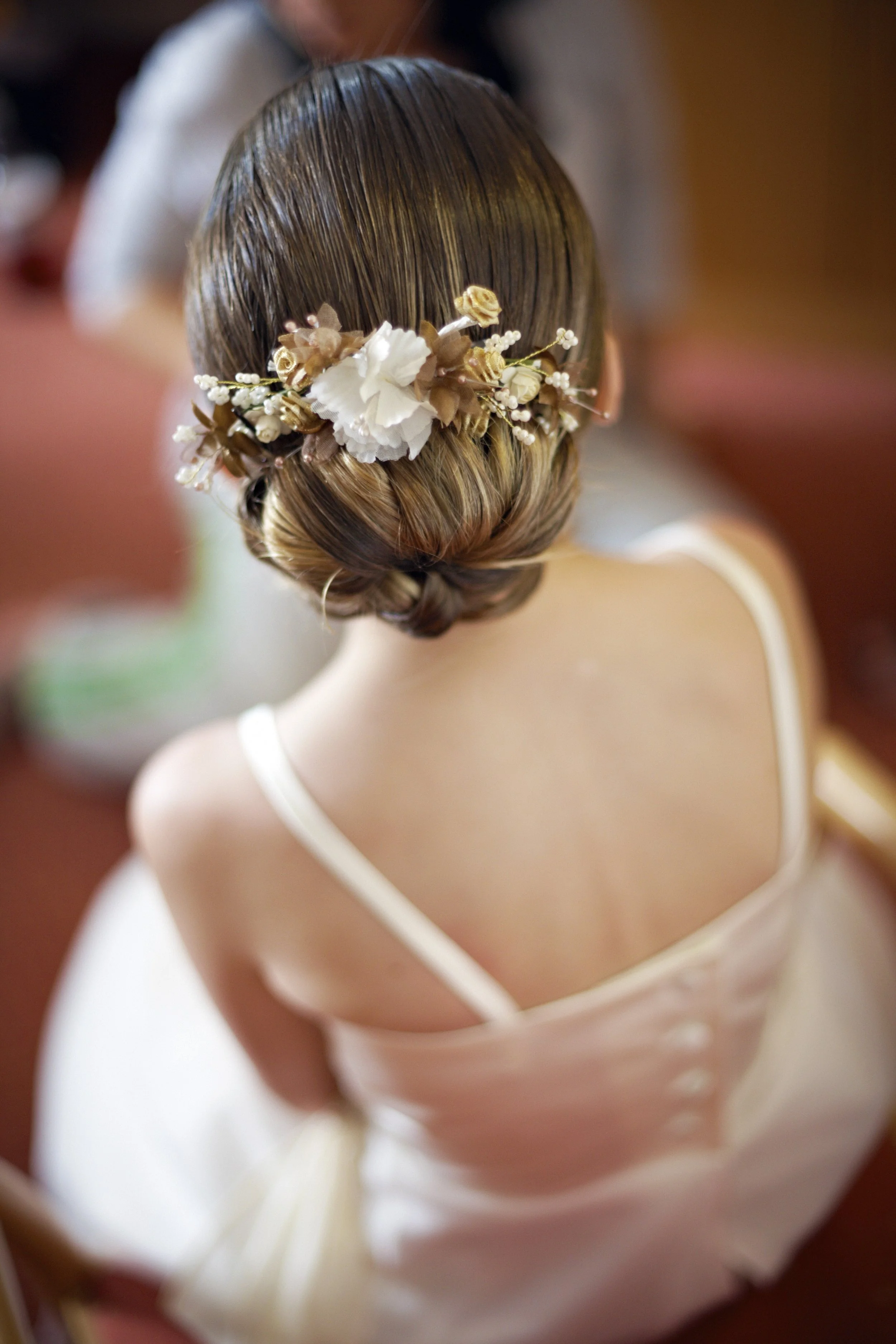Back view of a woman with brown hair styled in a low bun adorned with a floral hair accessory, wearing a white dress with spaghetti straps.