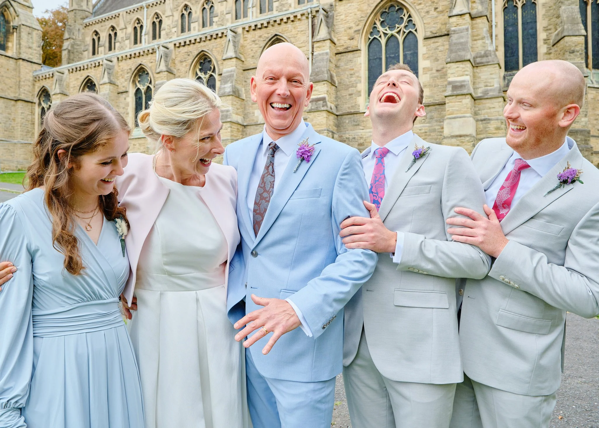 A group of six people dressed in formal attire, laughing and embracing each other in front of a stone church building.