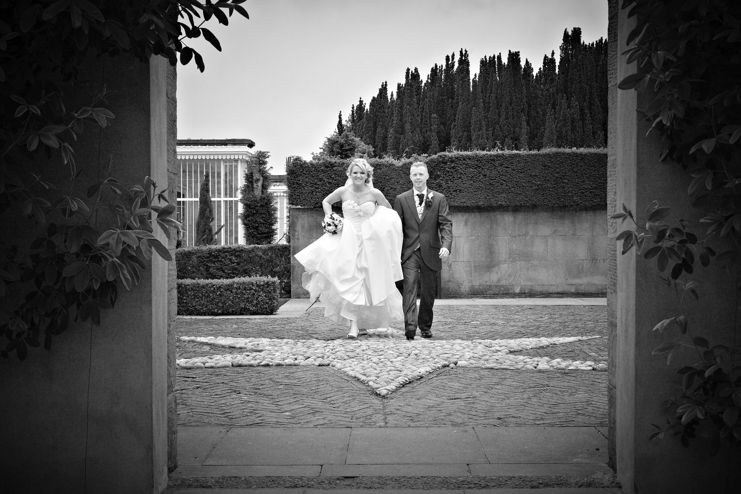 A bride and groom walking together outdoors, with the bride wearing a strapless wedding dress and holding a bouquet, the groom in a suit with a boutonniere. The scene is framed by an archway with foliage on either side, and the background shows a hed