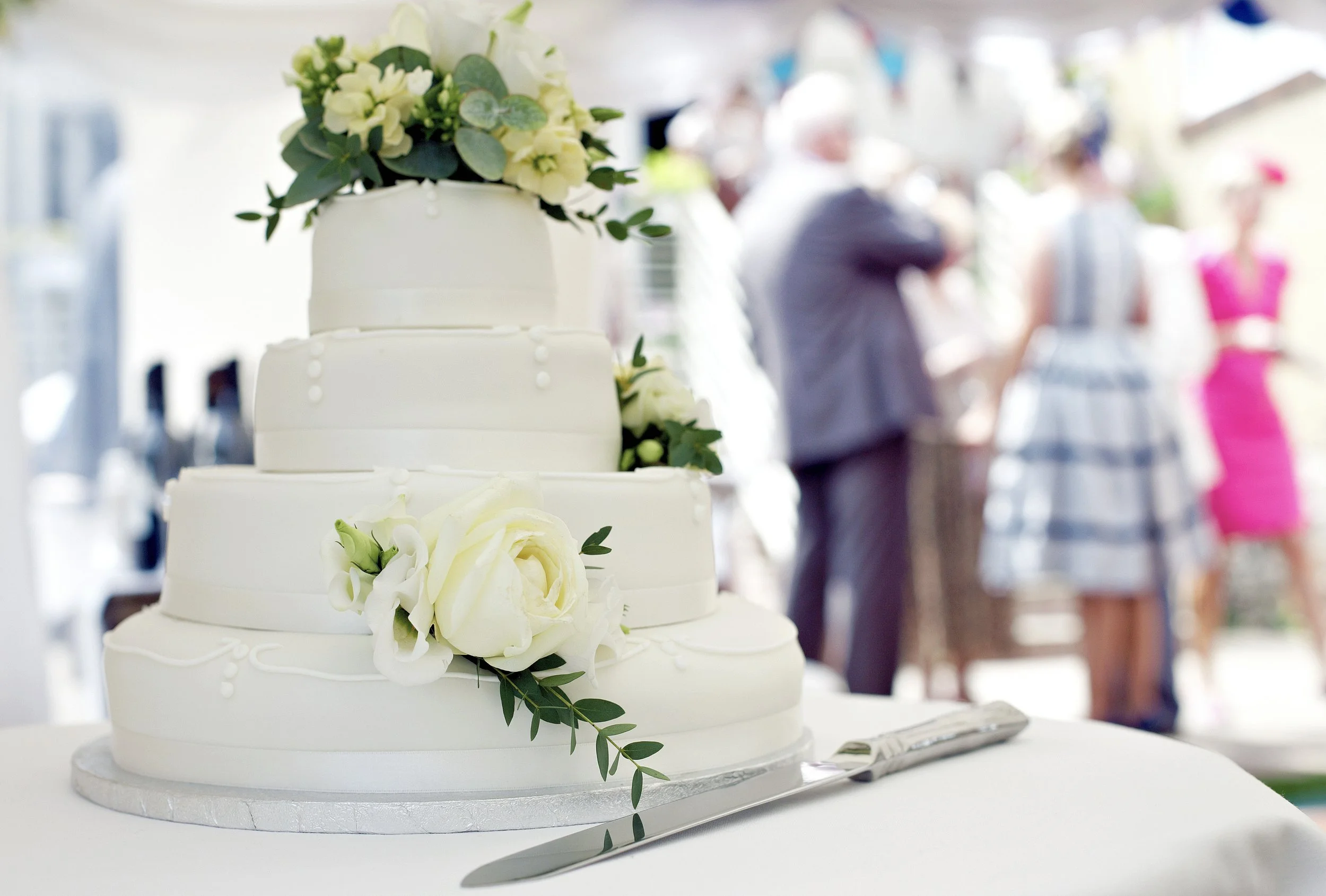 A white, tiered wedding cake decorated with white roses and green leaves, placed on a white table with a cake knife beside it, at a wedding reception.