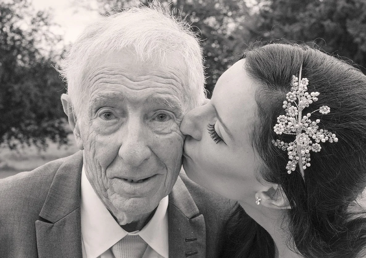 A young woman is giving an elderly man a kiss on the cheek outdoors, with trees in the background.