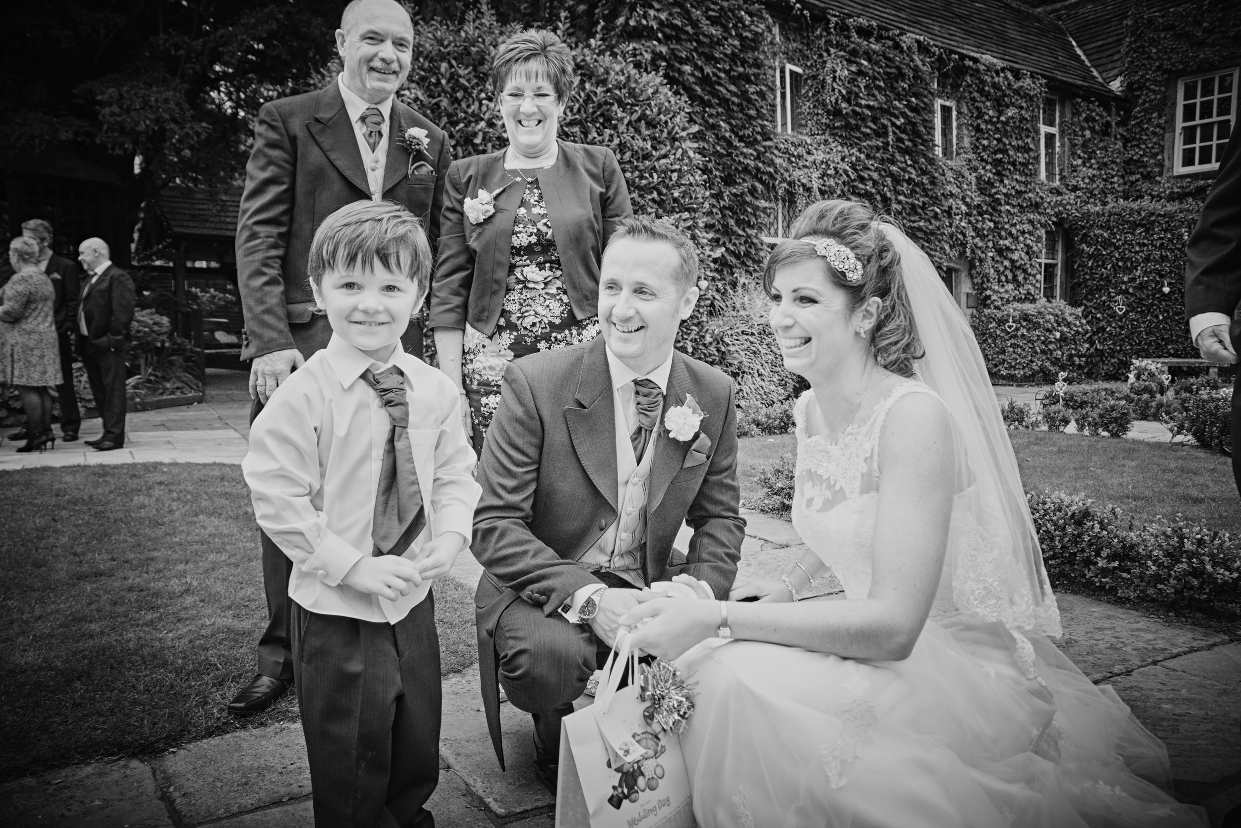 A wedding celebration in an outdoor garden with a group of people. The bride is smiling and holding a gift bag, and the groom, along with family members and friends, are gathered around, all smiling.