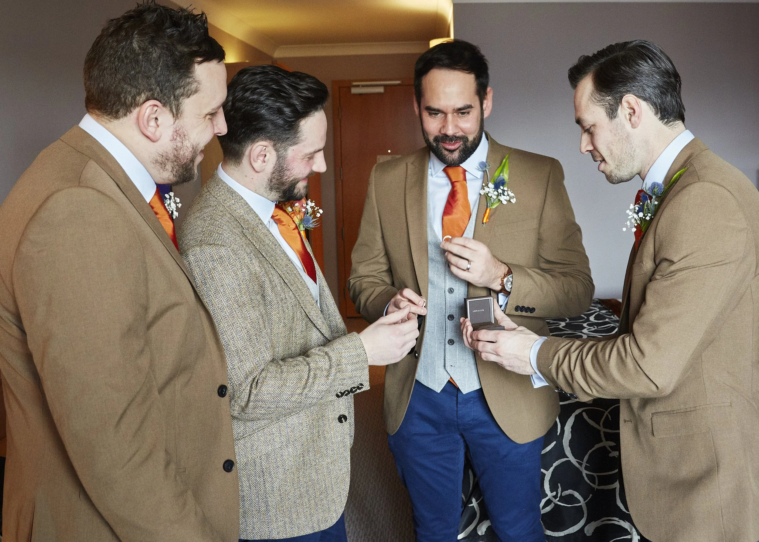 Five men dressed in suits standing in a circle, looking at each other, with one of the men holding an open ring box.