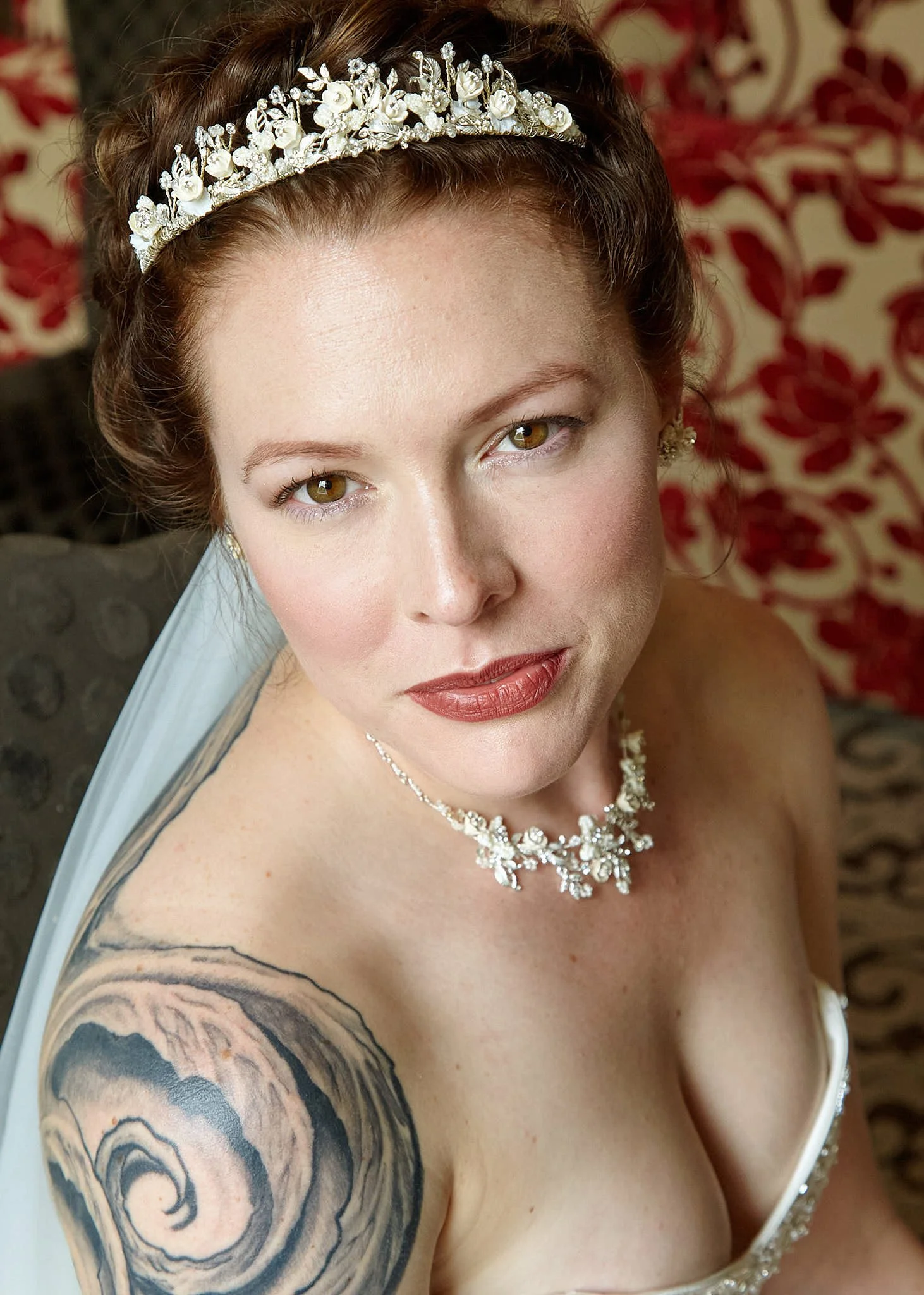 Close-up portrait of a woman with light skin, brown hair styled with a decorative tiara, wearing a pearl necklace and earrings, showing the right side of her face and shoulder with a tattoo of a rose, on a background with red and beige floral pattern