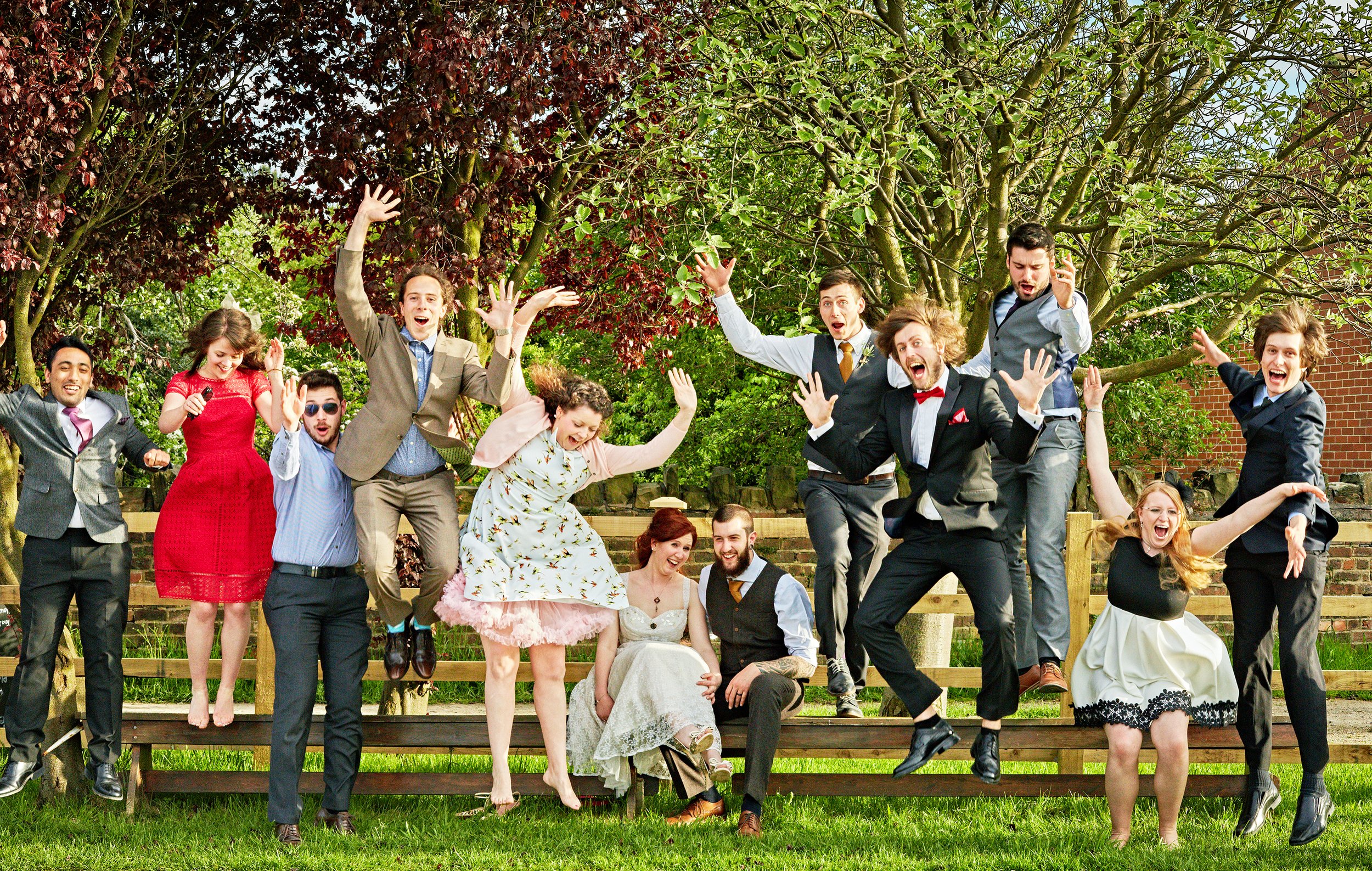 Group of people celebrating outdoors, some jumping, smiling, and making expressive gestures under trees.