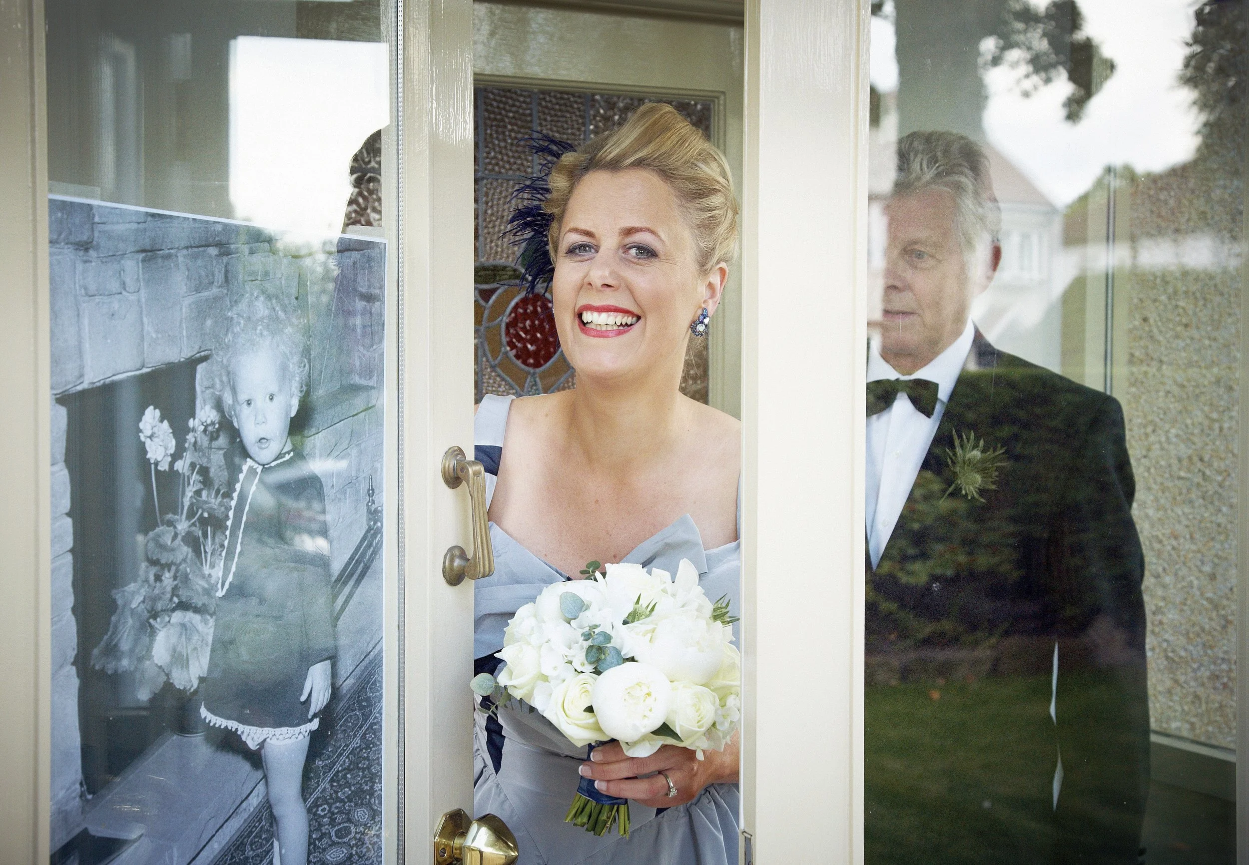 A woman in a wedding dress holding a bouquet of white flowers stands at the door of her home, smiling and looking outside, as an older man in a tuxedo looks on behind the glass door.