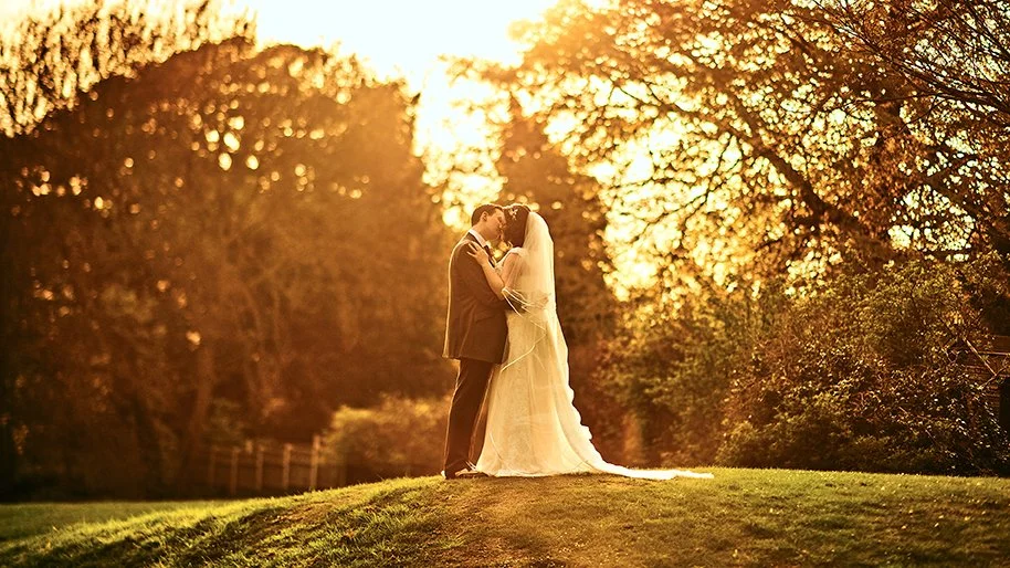 A bride and groom kissing on a grassy hill during sunset with trees in the background.