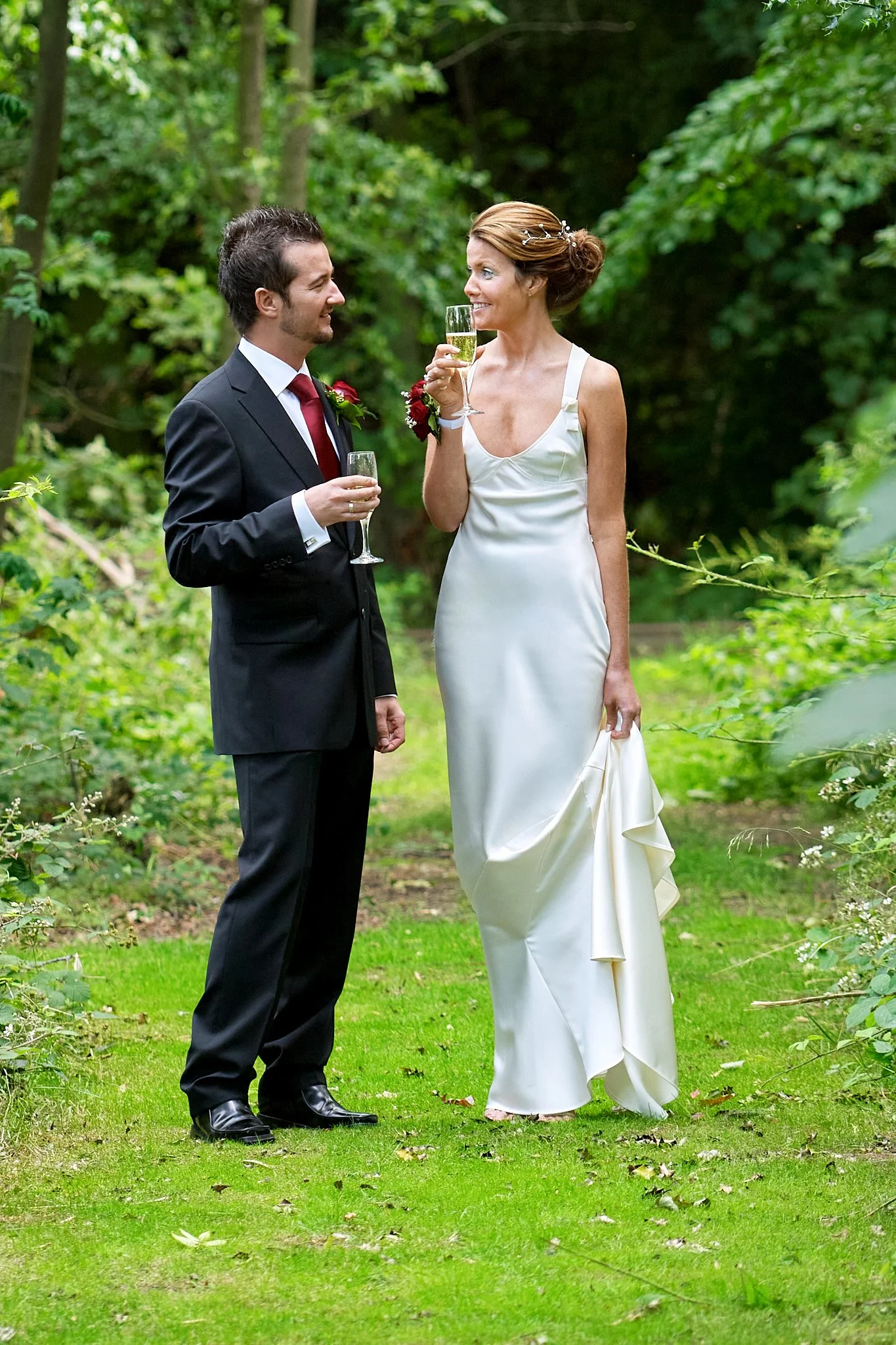 A man and woman in wedding attire standing outdoors, holding glasses of champagne and smiling at each other.