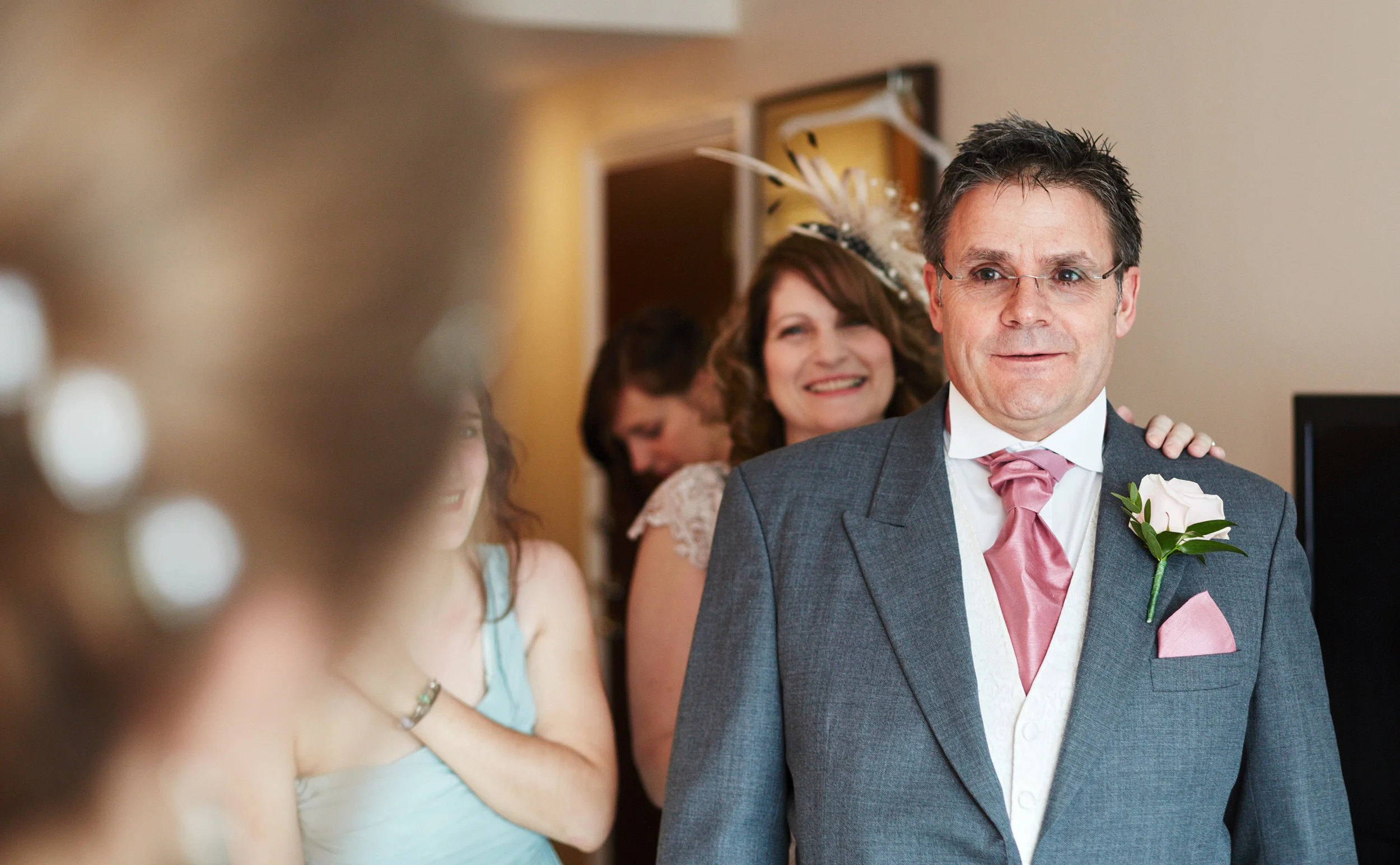 A man in a suit with a pink tie and boutonniere is smiling, standing close to a woman wearing a feathered hat, with several women and a young girl in the background at a social gathering or celebration.
