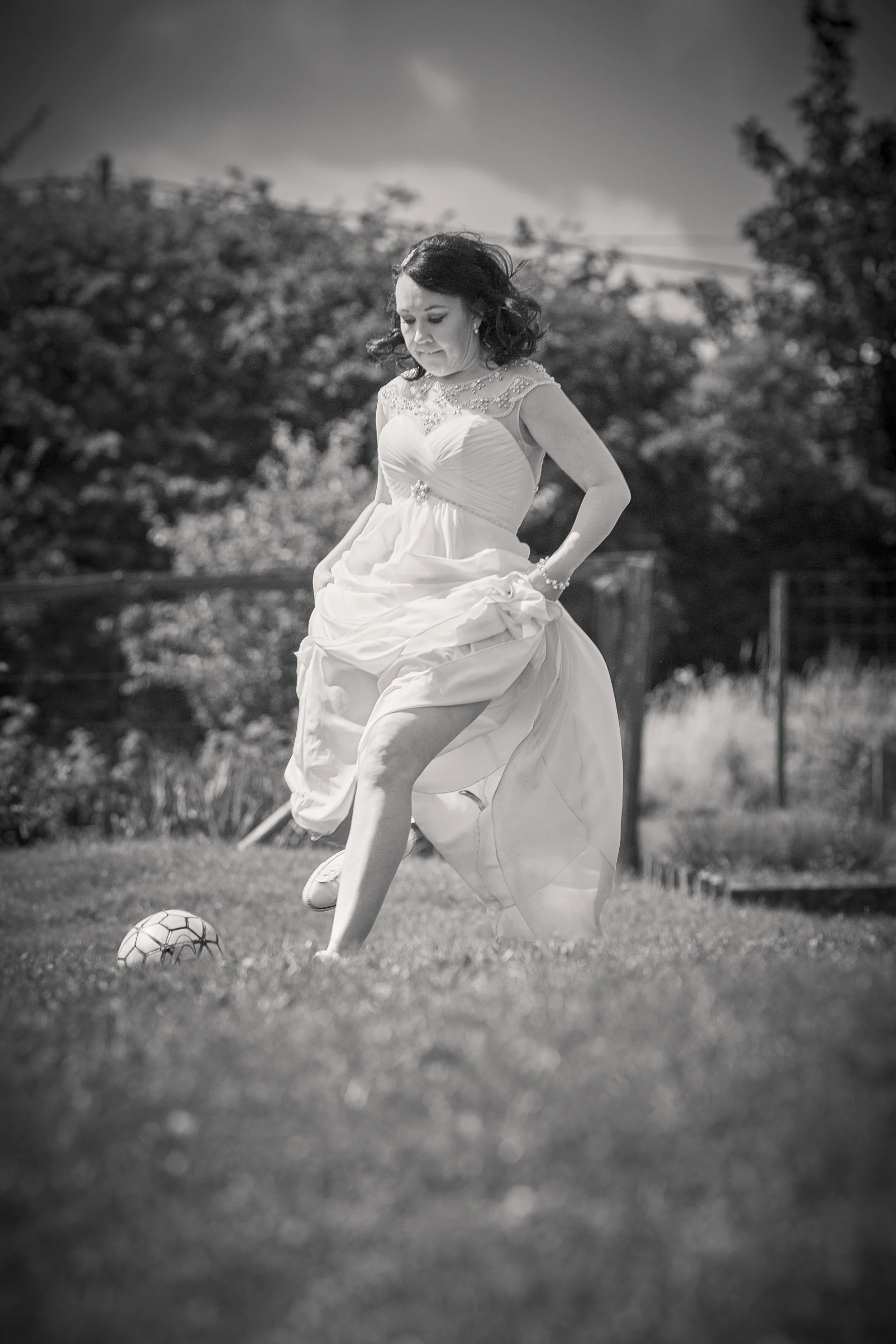 A woman in a wedding dress playing soccer outdoors on grass, with trees and fencing in the background, in black and white.