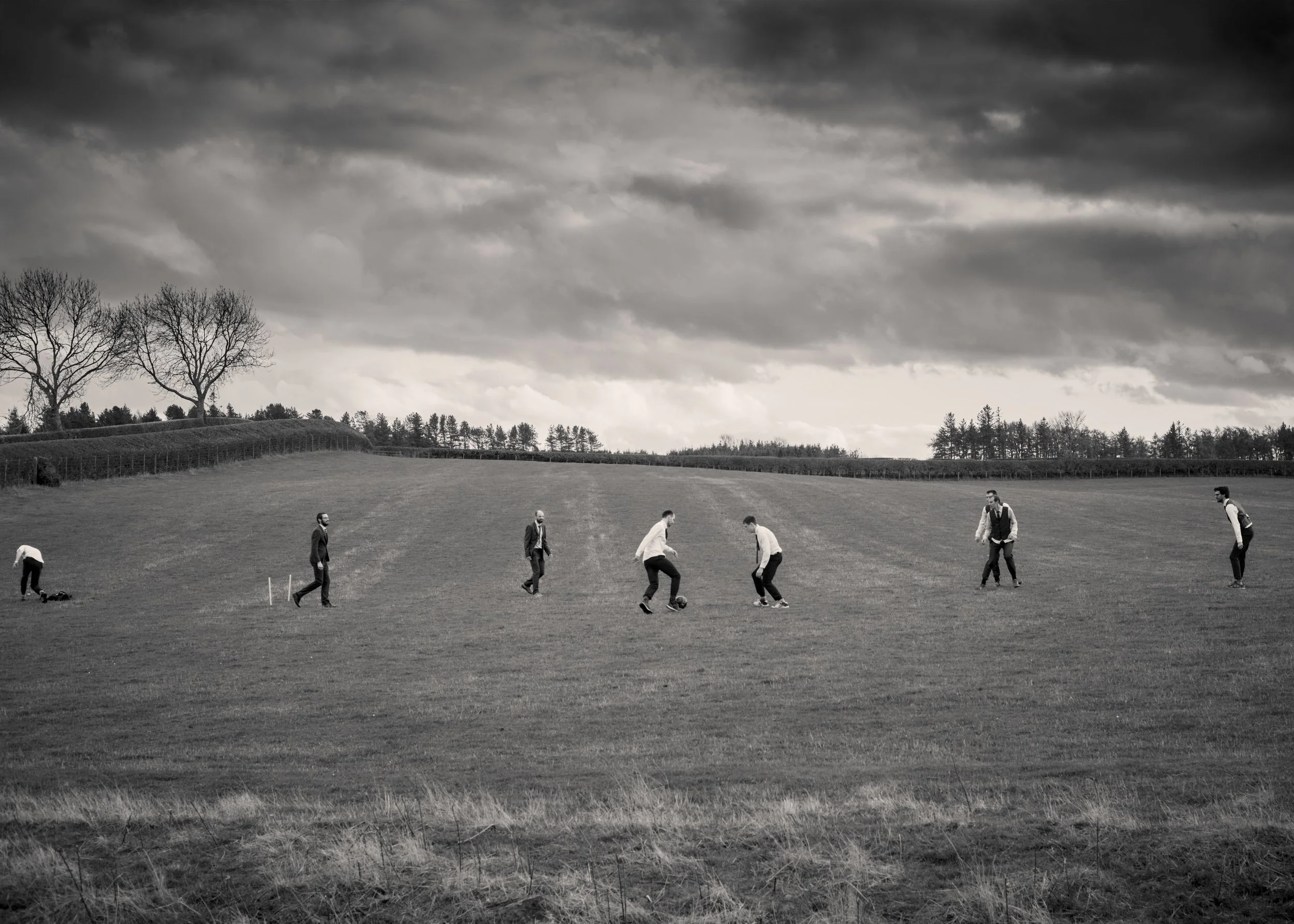 Black and white landscape with cloudy sky, trees, and people playing golf on an open field.