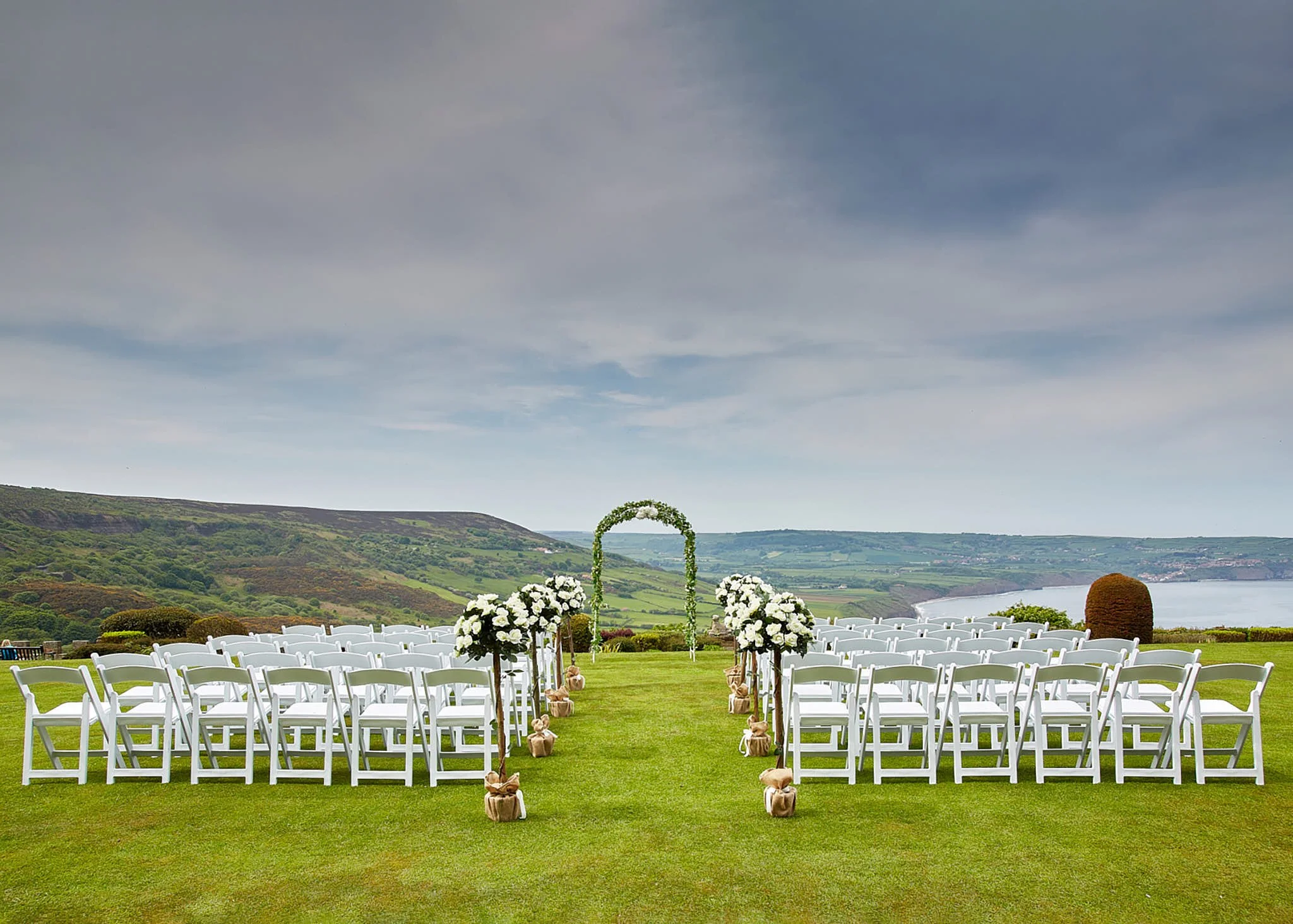 Outdoor wedding ceremony setup with white chairs arranged in rows on green grass, decorated with white flowers and small burlap-wrapped bundles along the aisle, overlooking a river and hills under a cloudy sky.