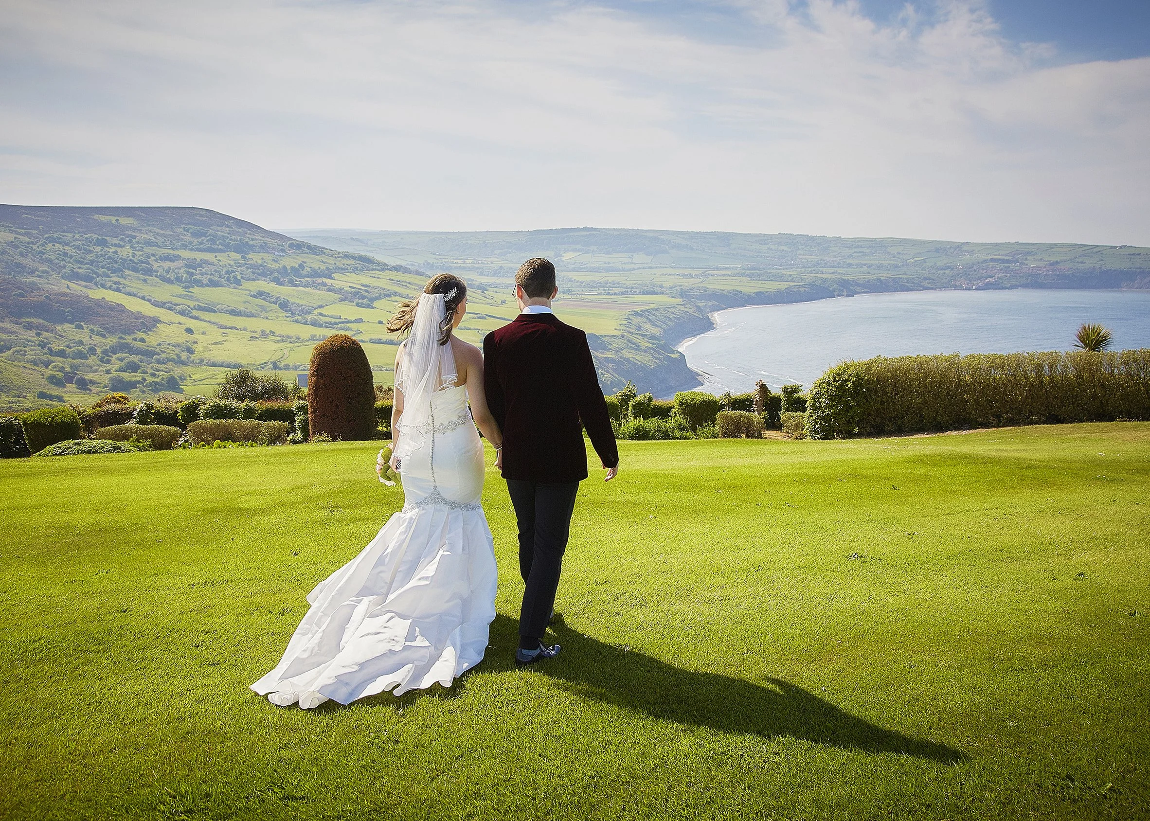 A bride and groom holding hands while walking on lush green grass overlooking the ocean and rolling hills on a partly cloudy day.