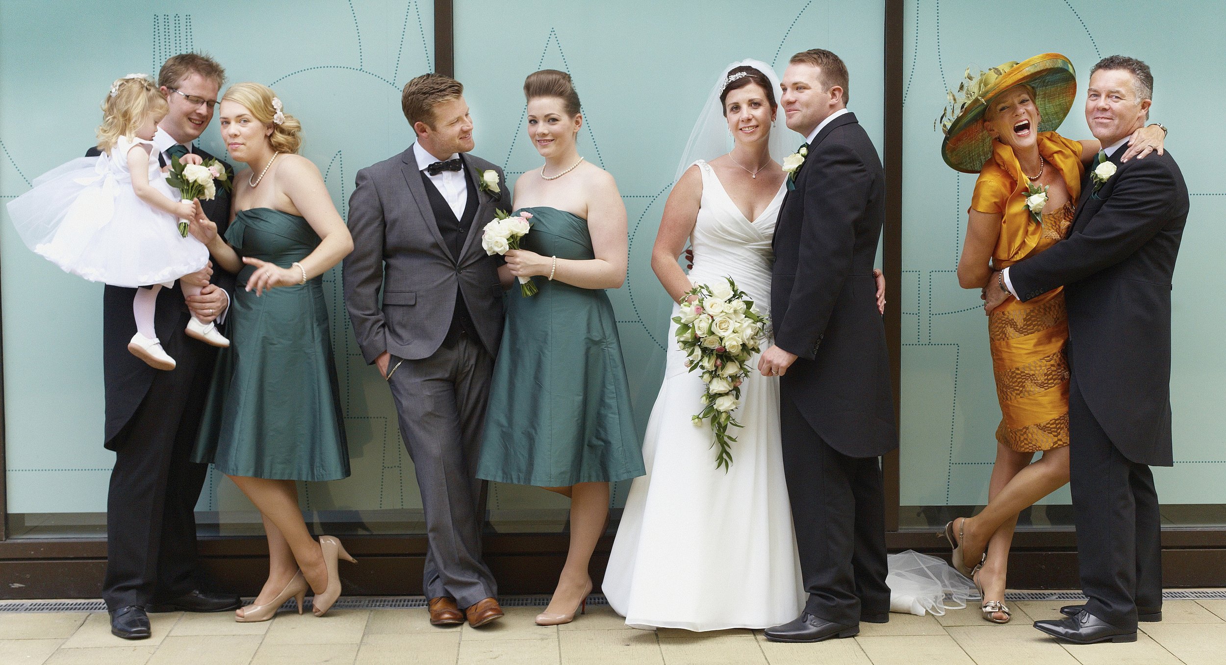 Group of people at a wedding, with brides and grooms, standing in front of a light green background with patterns.