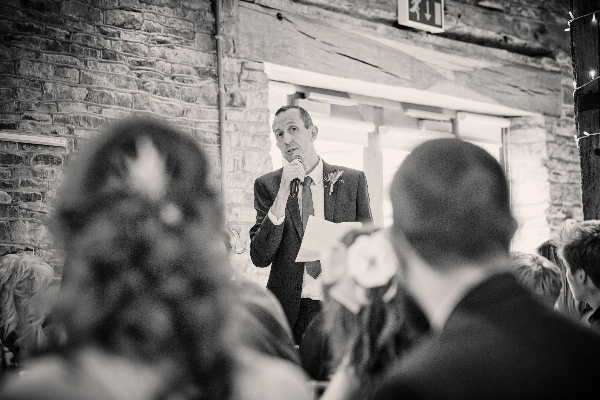 A man in a suit holding a microphone and reading from a paper, speaking to an audience in a rustic, brick-walled room with exposed wooden beams.