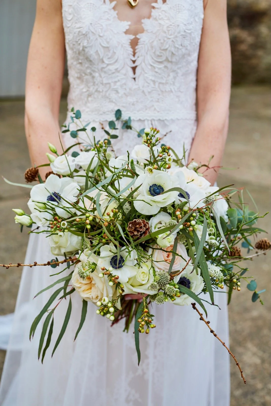 A bride in a white lace dress holding a bouquet of white flowers, greenery, and pinecones.