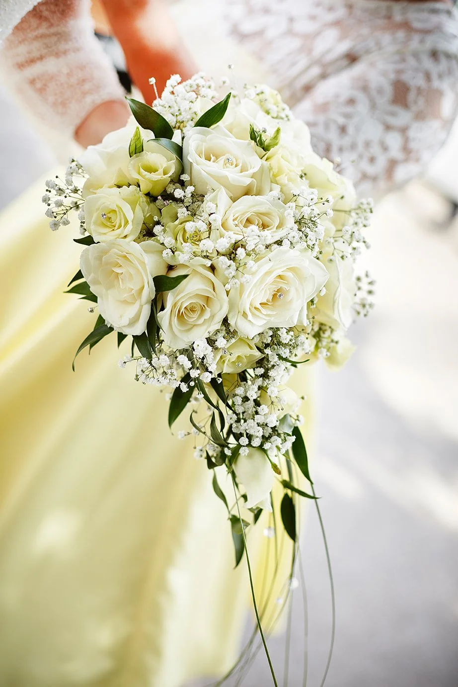 A bouquet of white roses, baby's breath, and greenery held by a person wearing a lace dress and yellow skirt, likely at a wedding.