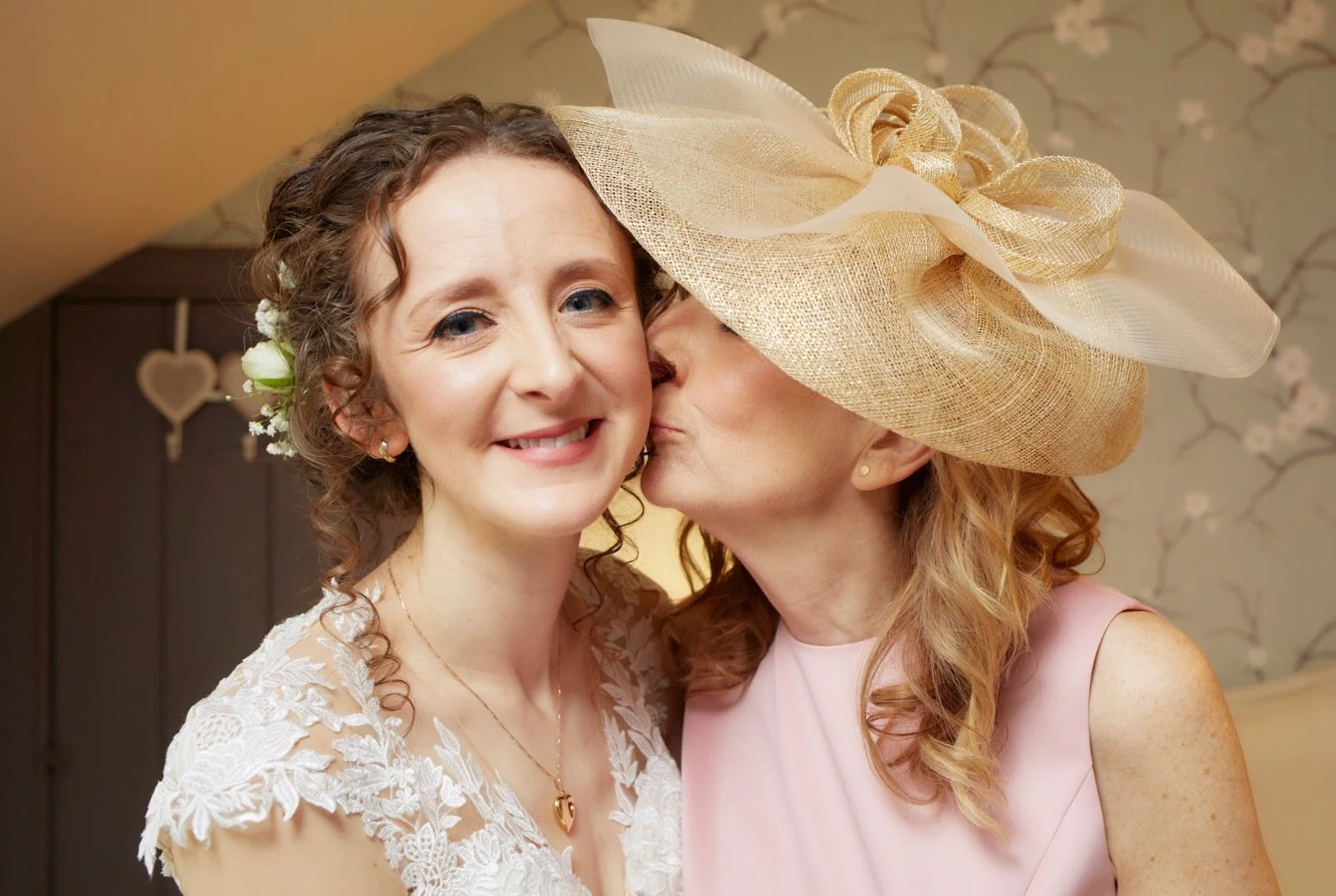 A woman with curly hair in a lace dress is being kissed on the cheek by another woman wearing a large beige hat, in a room with floral wallpaper.