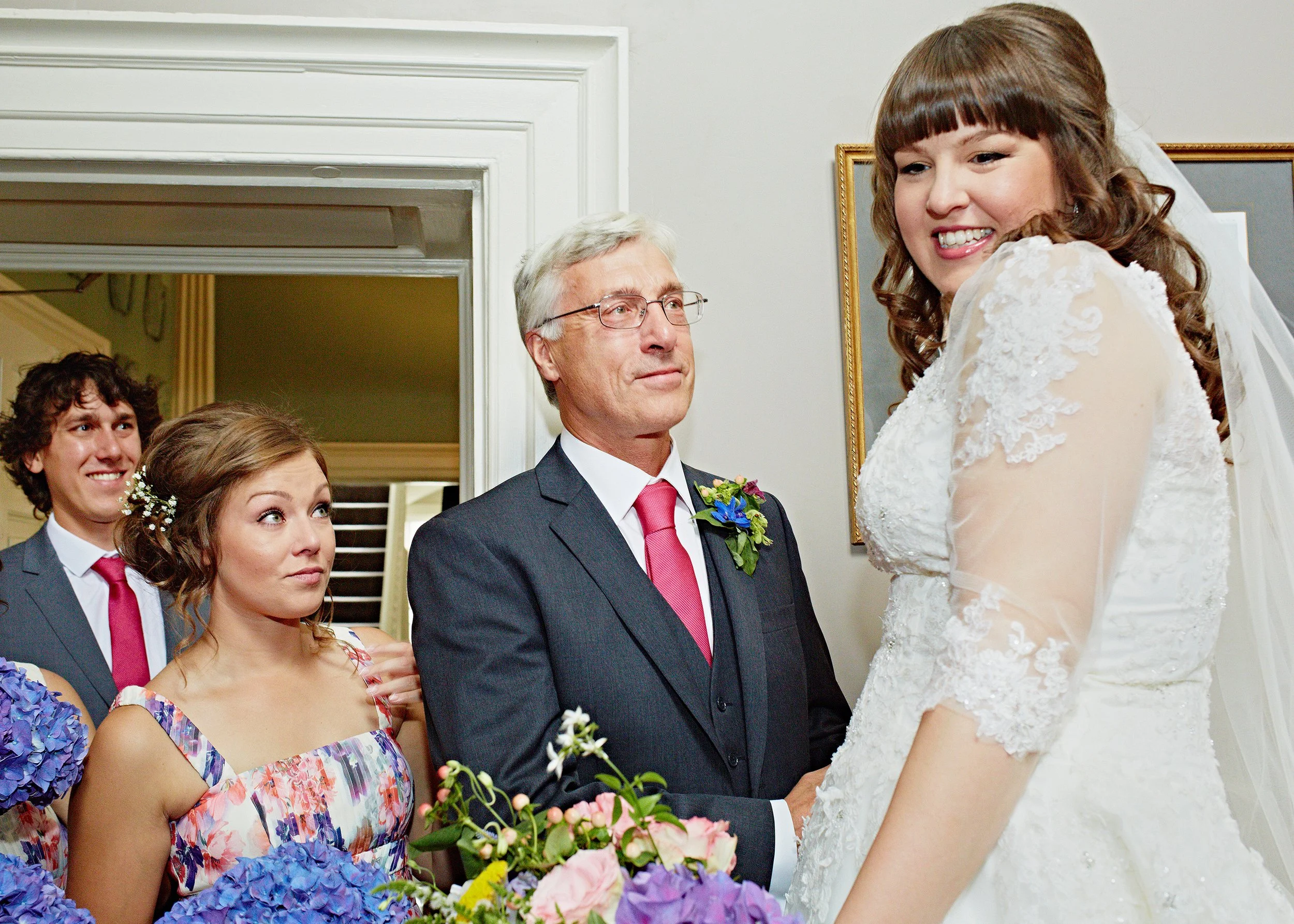 A bride and groom exchanging vows during a wedding ceremony, surrounded by wedding party members holding flowers.